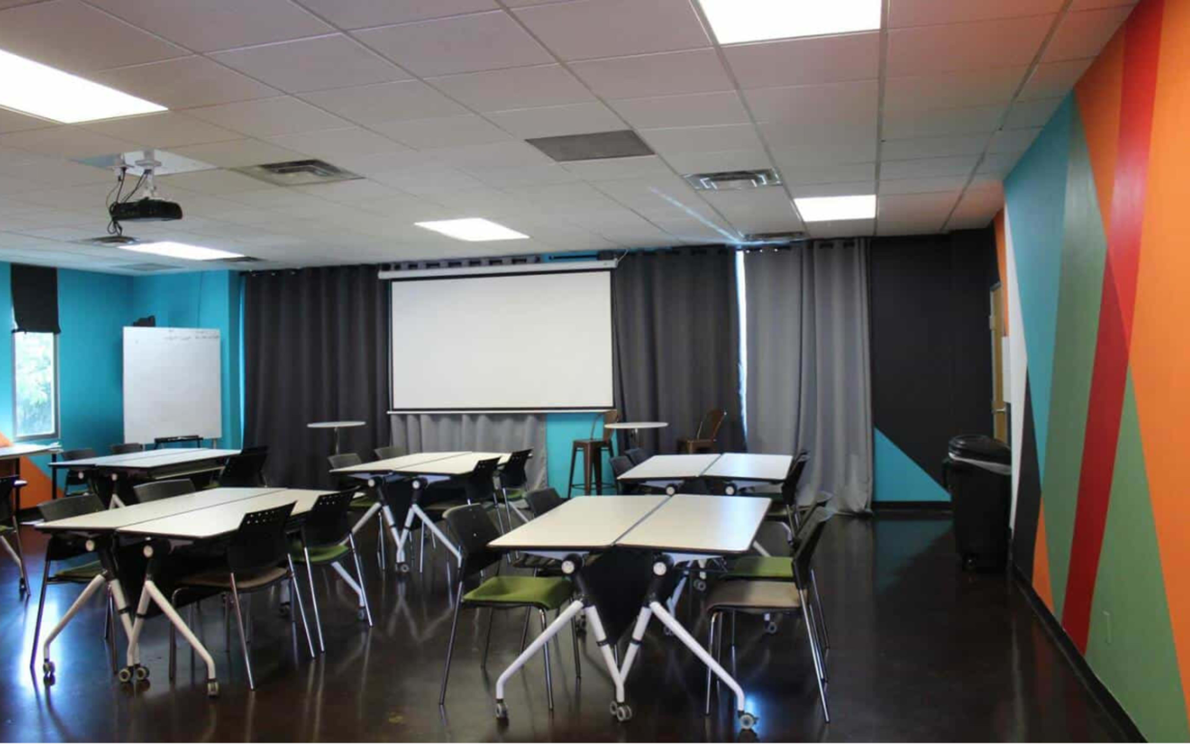 A modern classroom setup with several tables and chairs arranged facing a projector screen, surrounded by colorful walls and curtains.