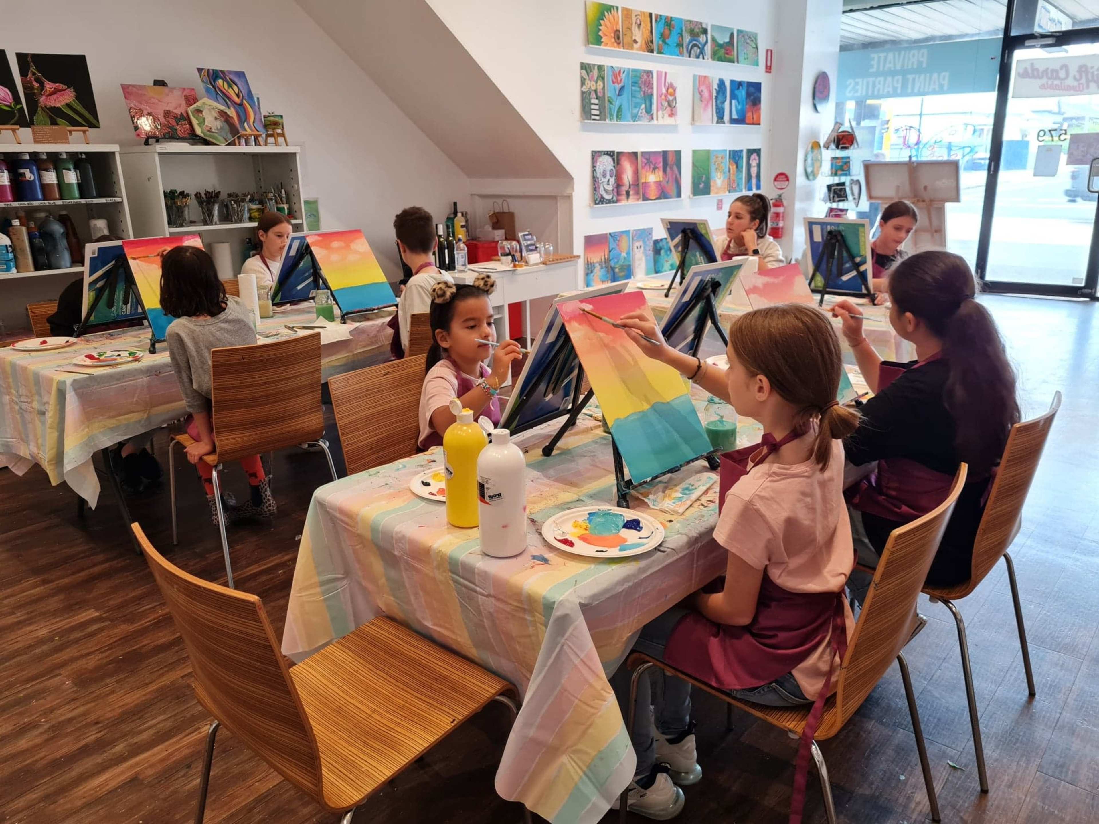 A group of children is painting on canvases at tables in a brightly lit art studio.