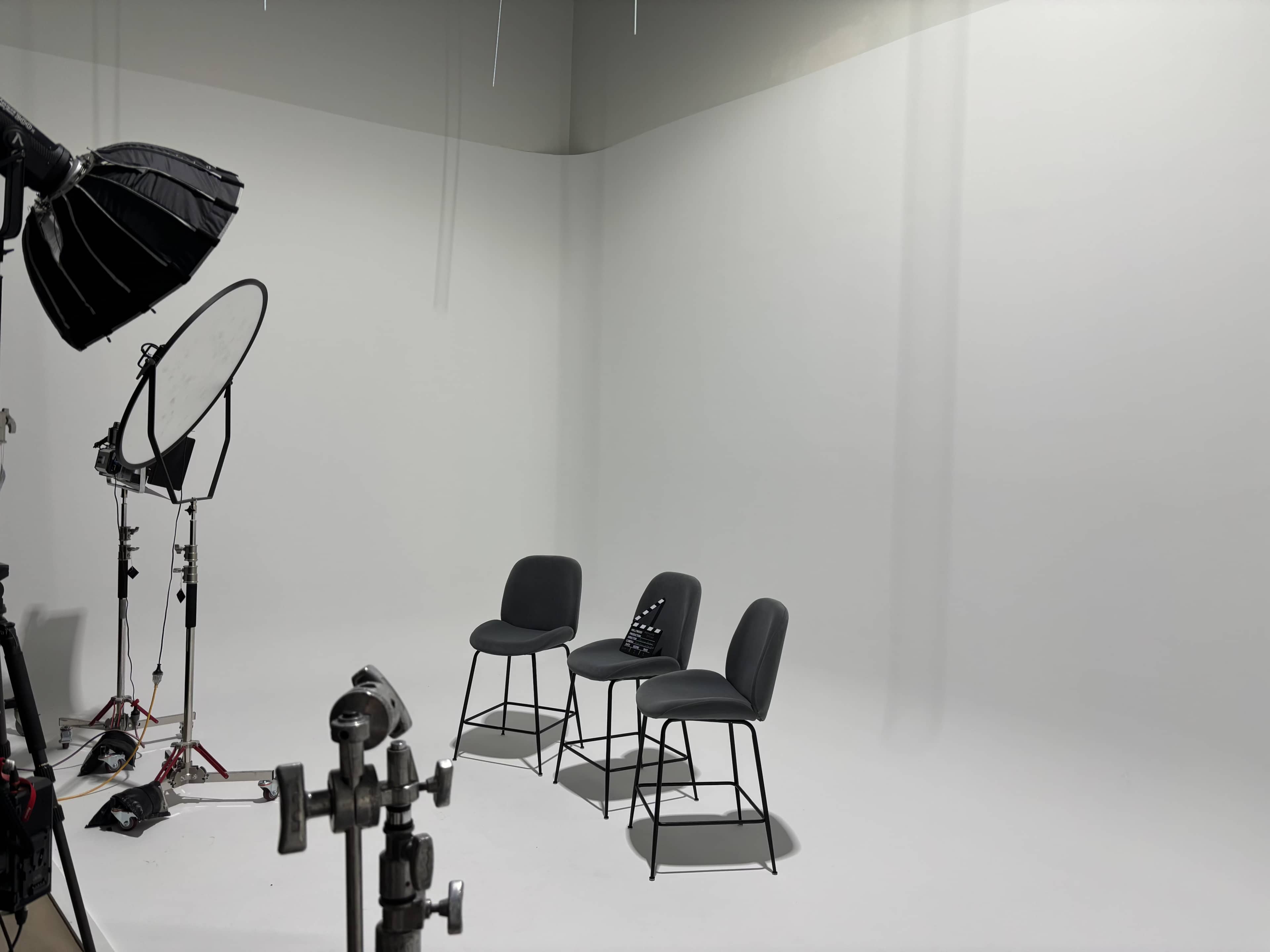 A photography studio is shown with three gray chairs positioned in front of a plain white backdrop, accompanied by studio lighting equipment.