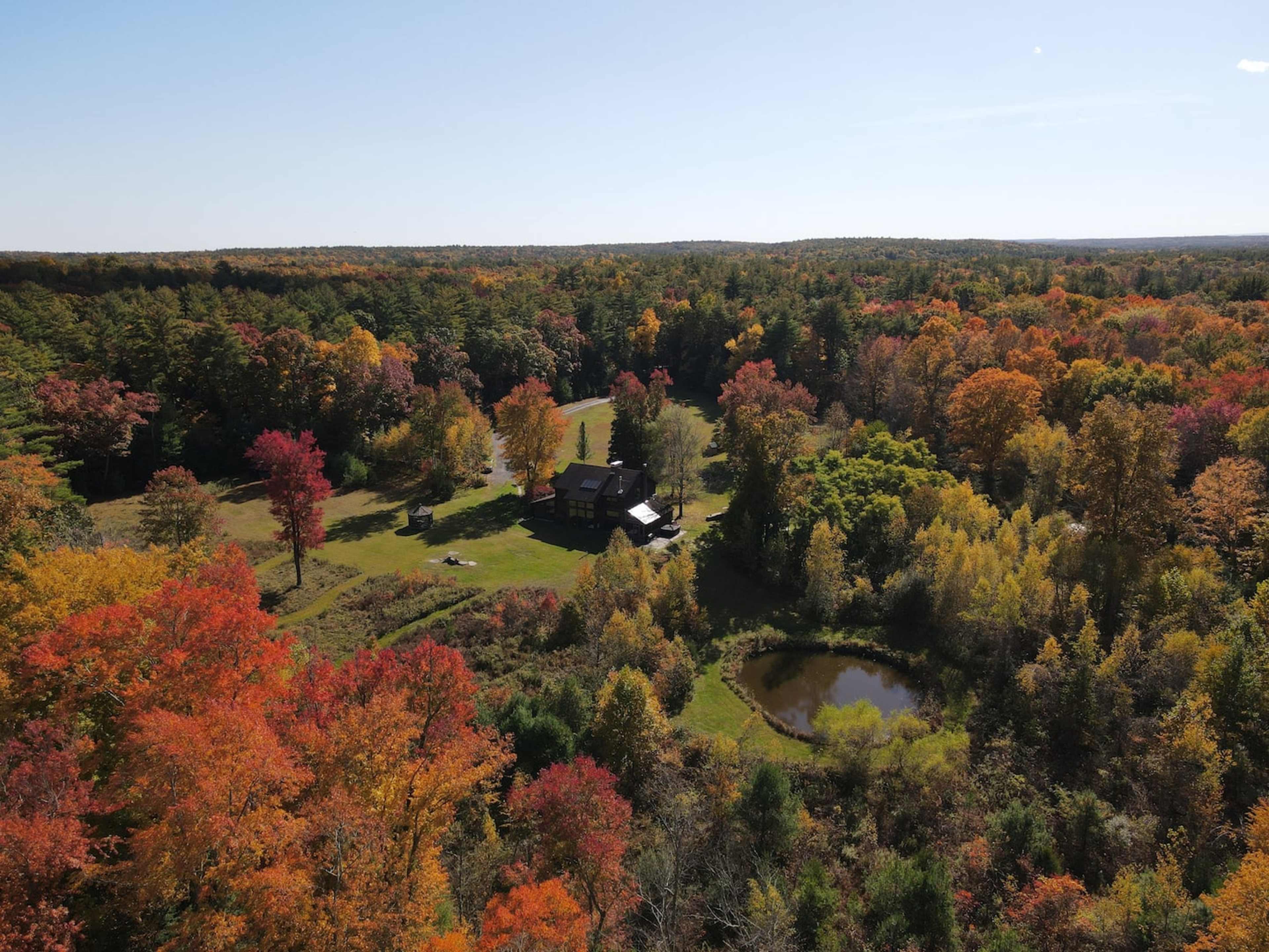 An aerial view captures a house surrounded by colorful autumn foliage and a small pond in a wooded landscape.