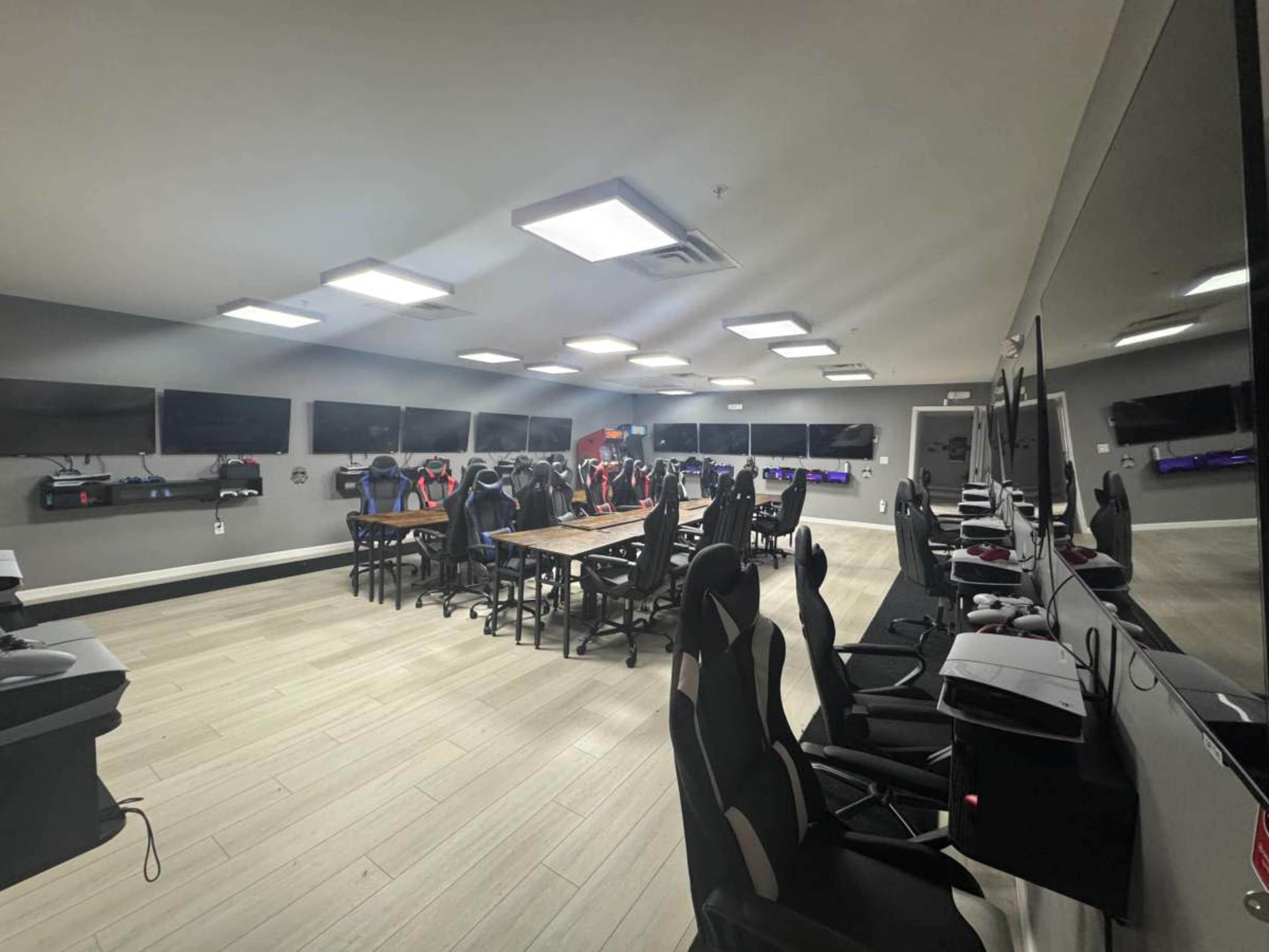 The image shows a modern gaming room with multiple black and colored gaming chairs arranged around a large table, and several screens mounted on the walls.