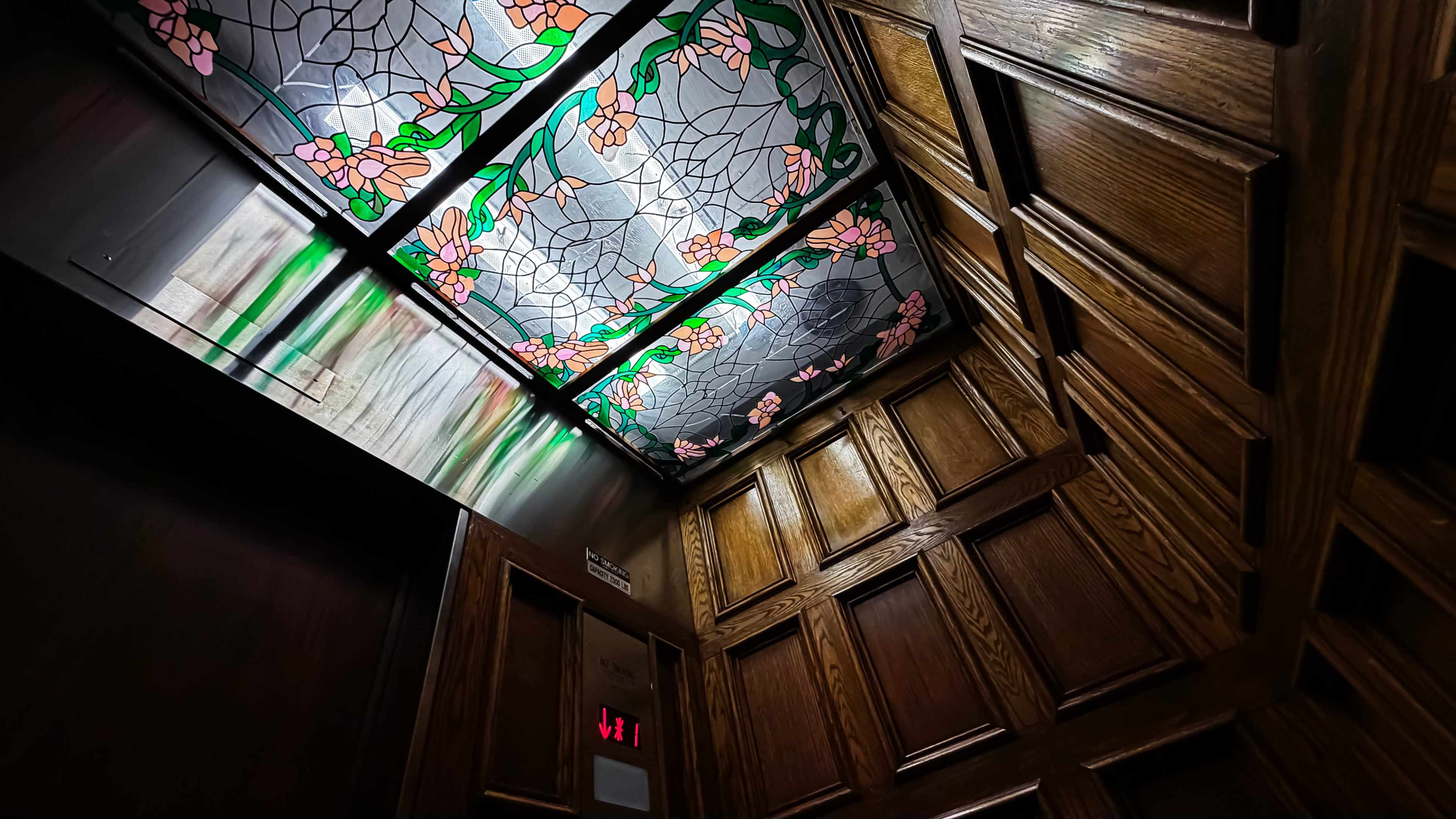 The image shows the interior of an elevator with wooden paneling and a stained glass ceiling featuring floral designs.
