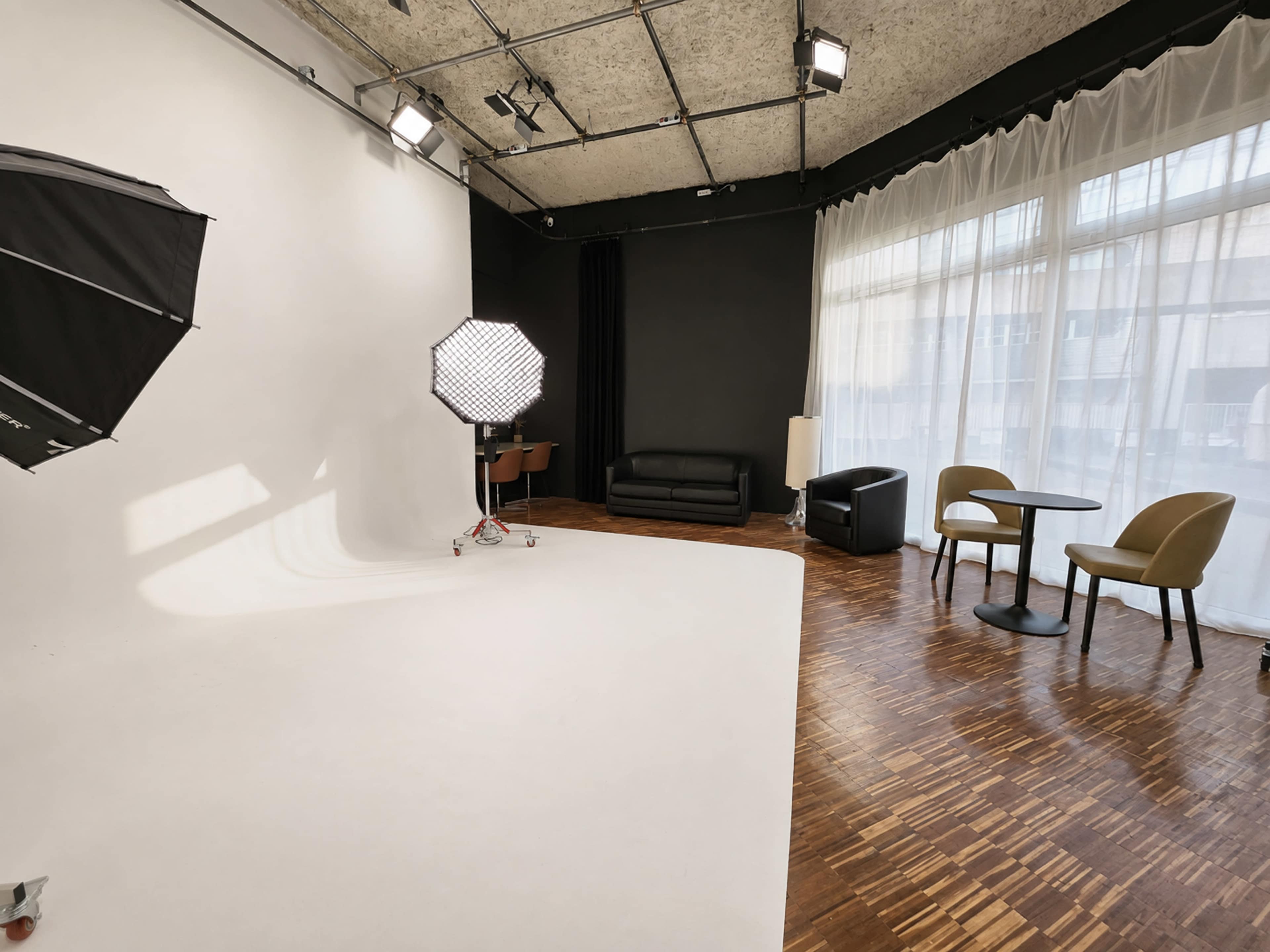 The image shows a photography studio with a white backdrop, lighting equipment, a black sofa, a black armchair, a round table, and large windows letting in natural light.