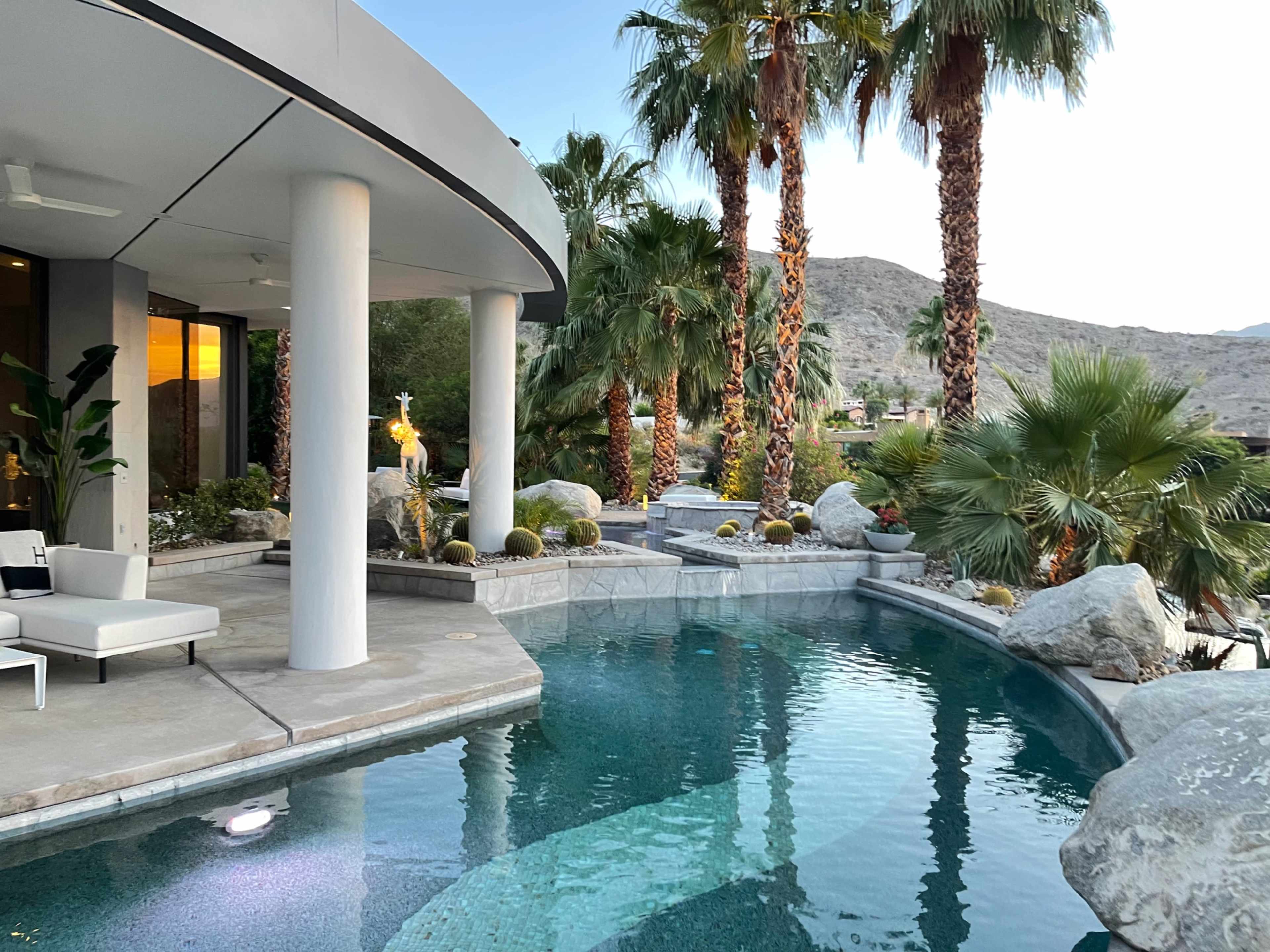 The scene features a modern patio with a curved pool surrounded by palm trees and rocky landscaping.