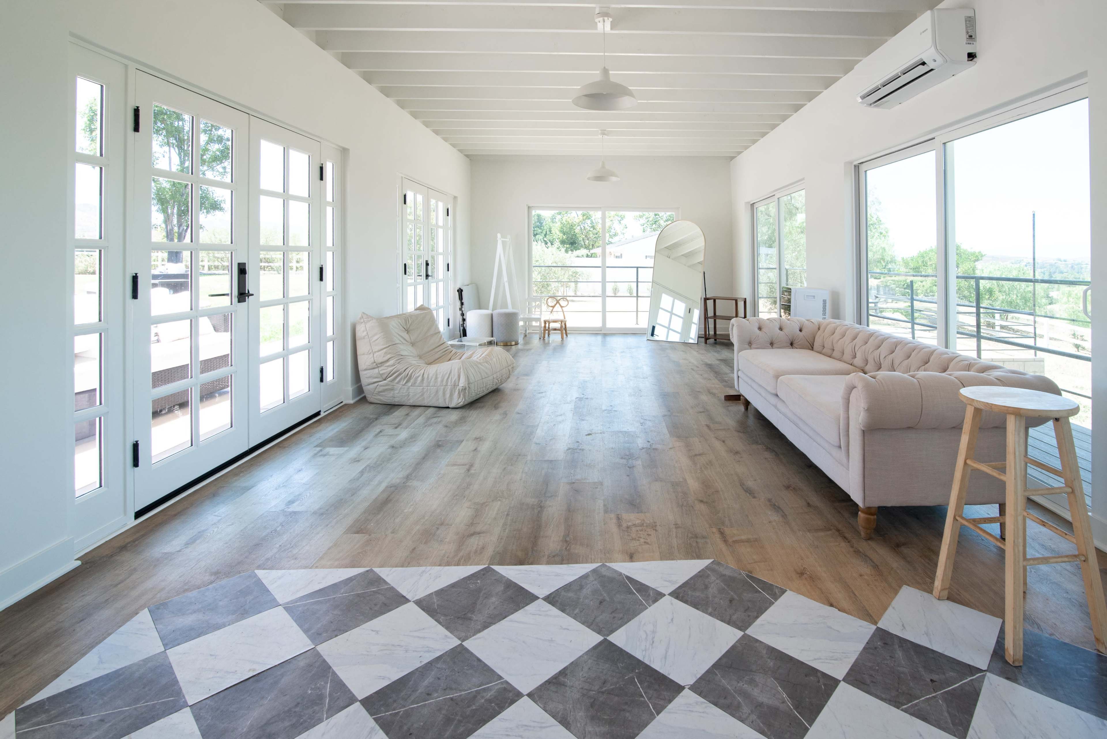 A spacious, well-lit interior features a beige sofa, a bean bag chair, and a patterned floor with large windows overlooking an outdoor area.