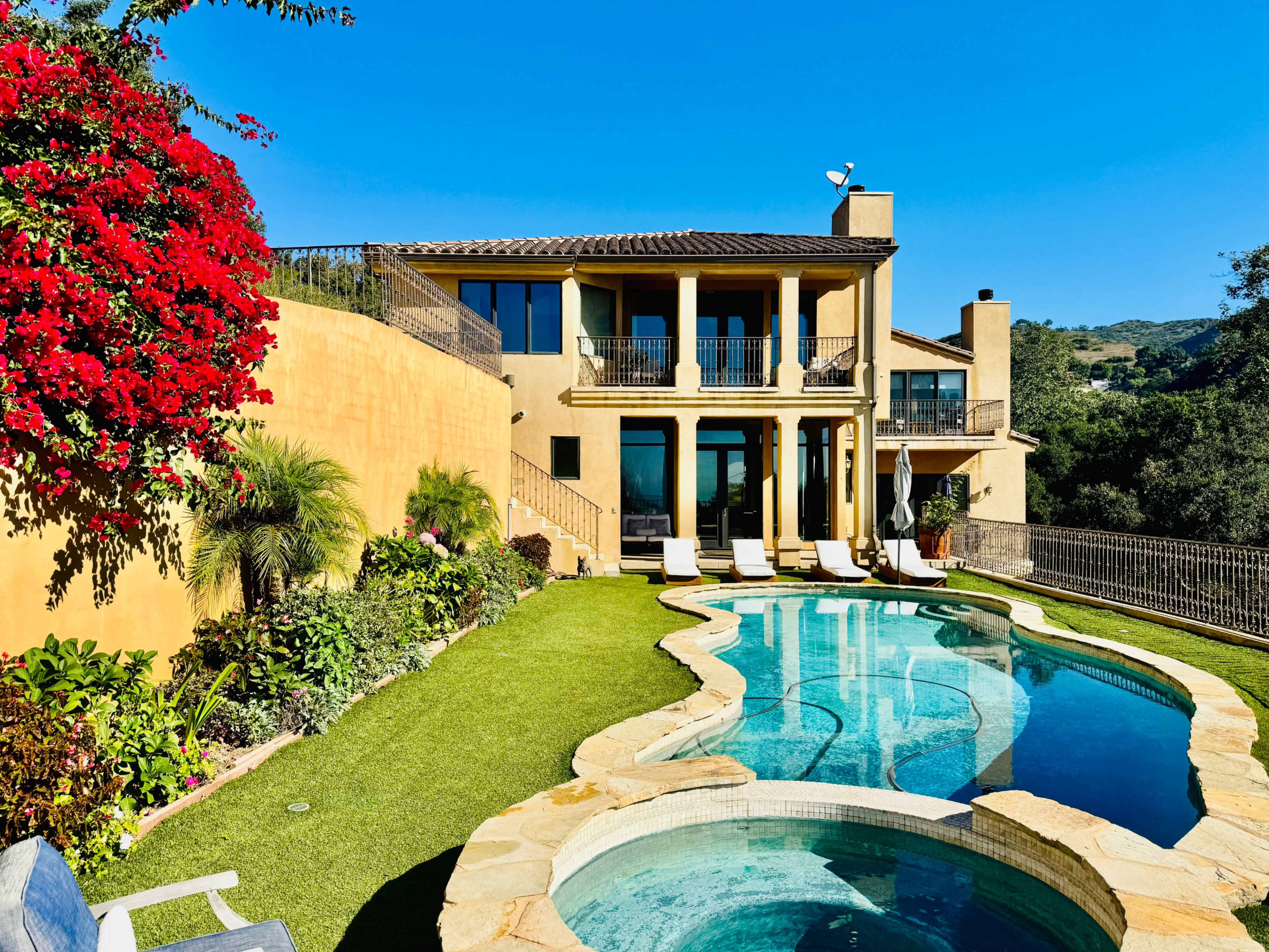 The image shows a well-maintained villa with a swimming pool and landscaped gardens, set against a clear blue sky.