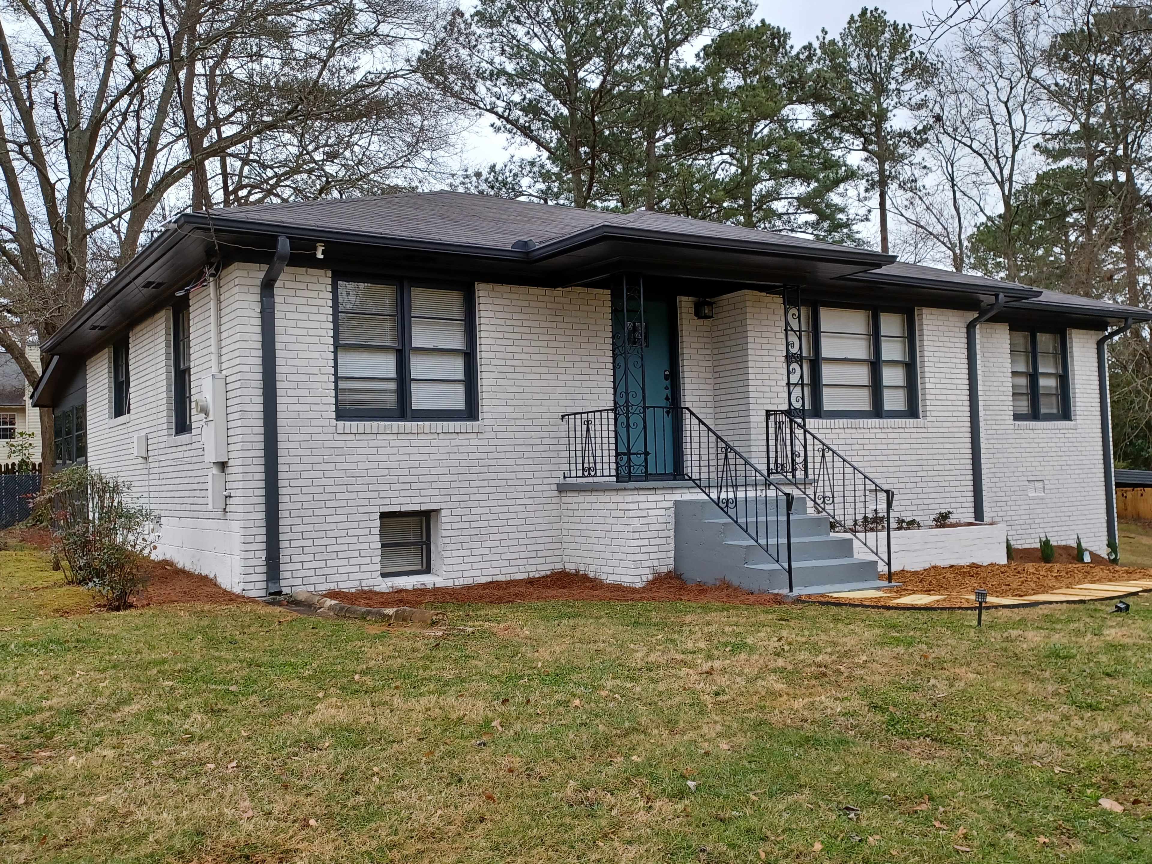 A single-story white brick house with black trim and stairs leads up to a teal front door, surrounded by green grass and trees.
