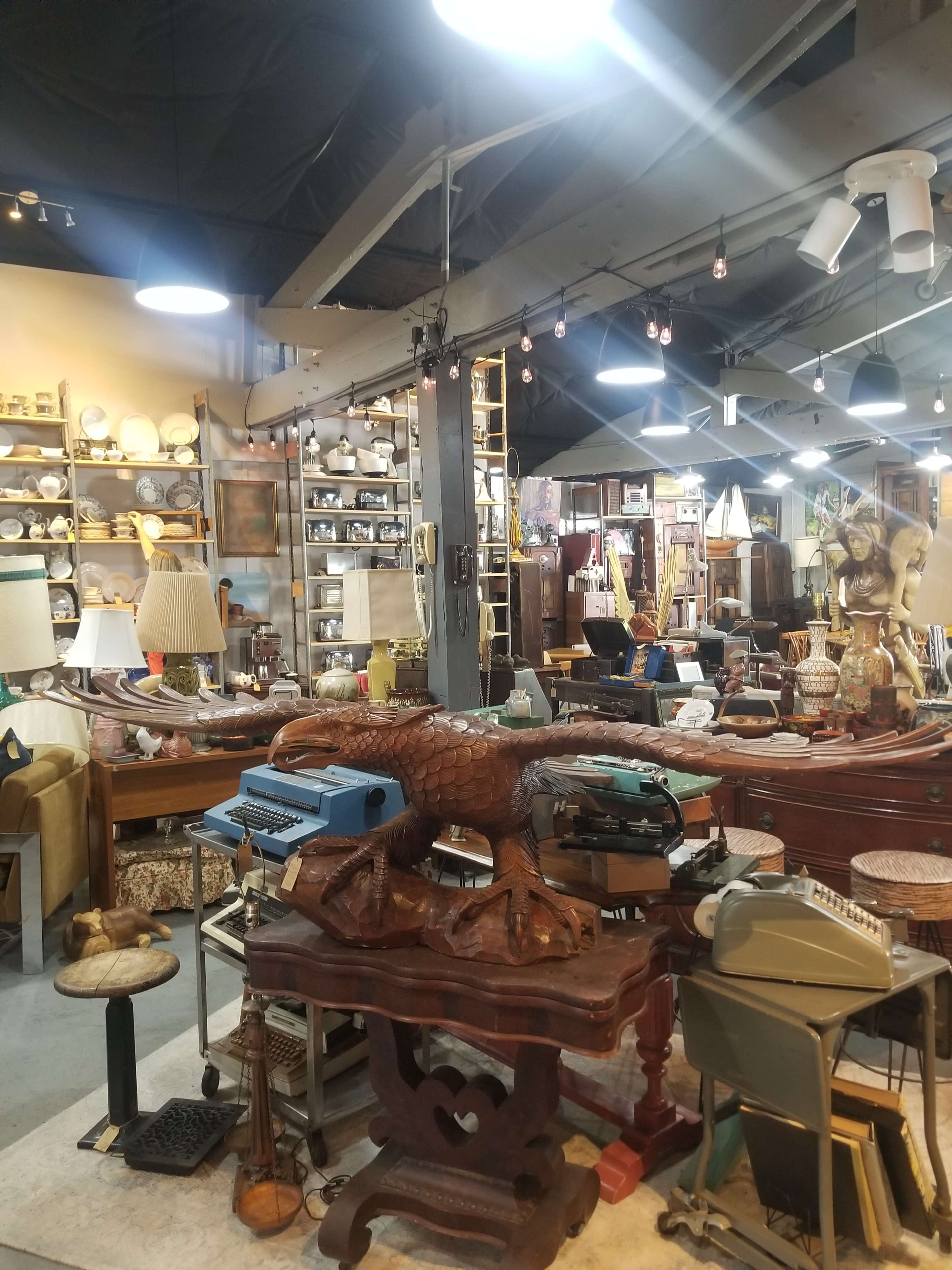 A cluttered antiques shop filled with a variety of vintage items, including furniture, lamps, and a typewriter, arranged across multiple displays.