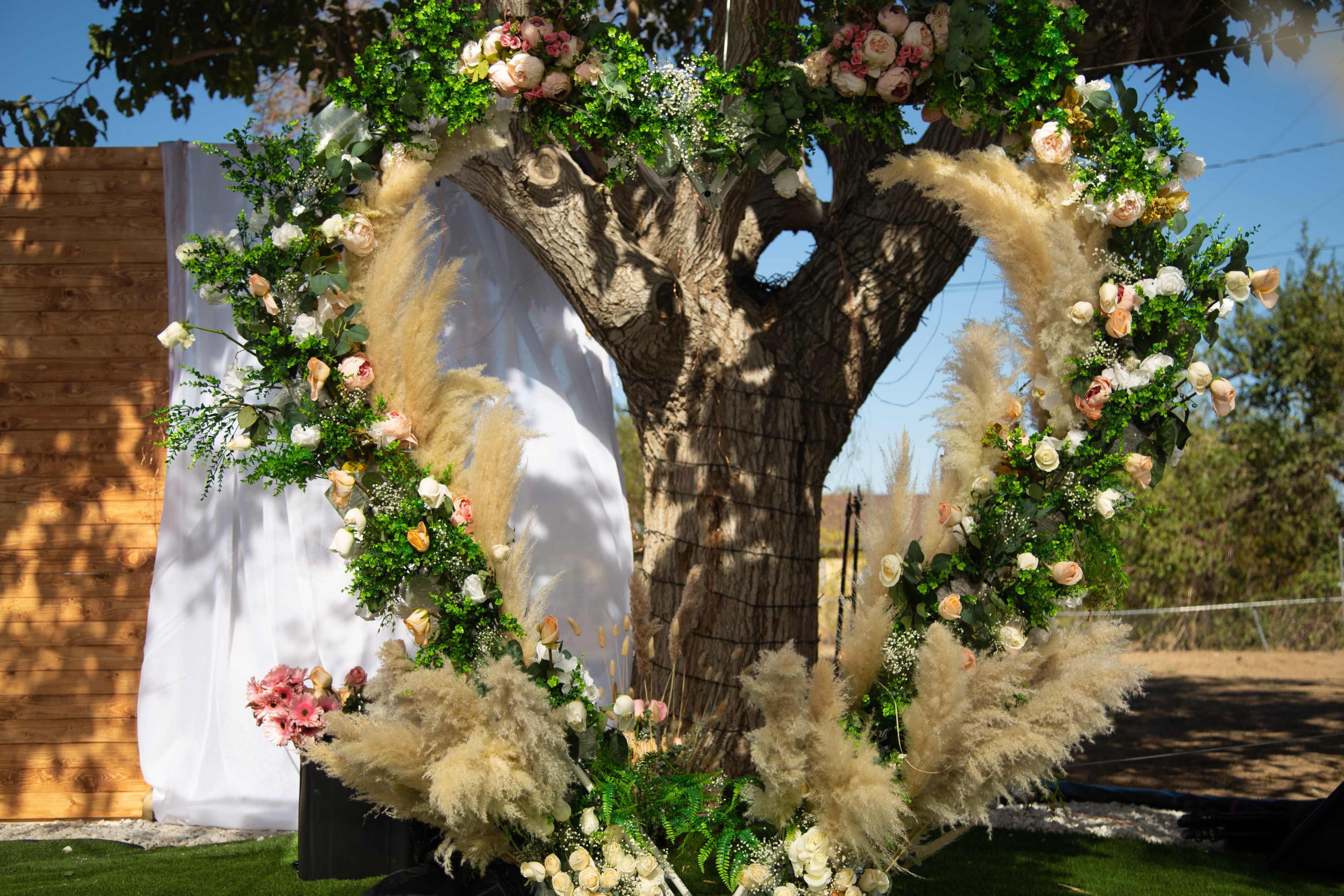 A large heart-shaped floral arrangement made of greenery, flowers, and pampas grass is positioned against a tree trunk.