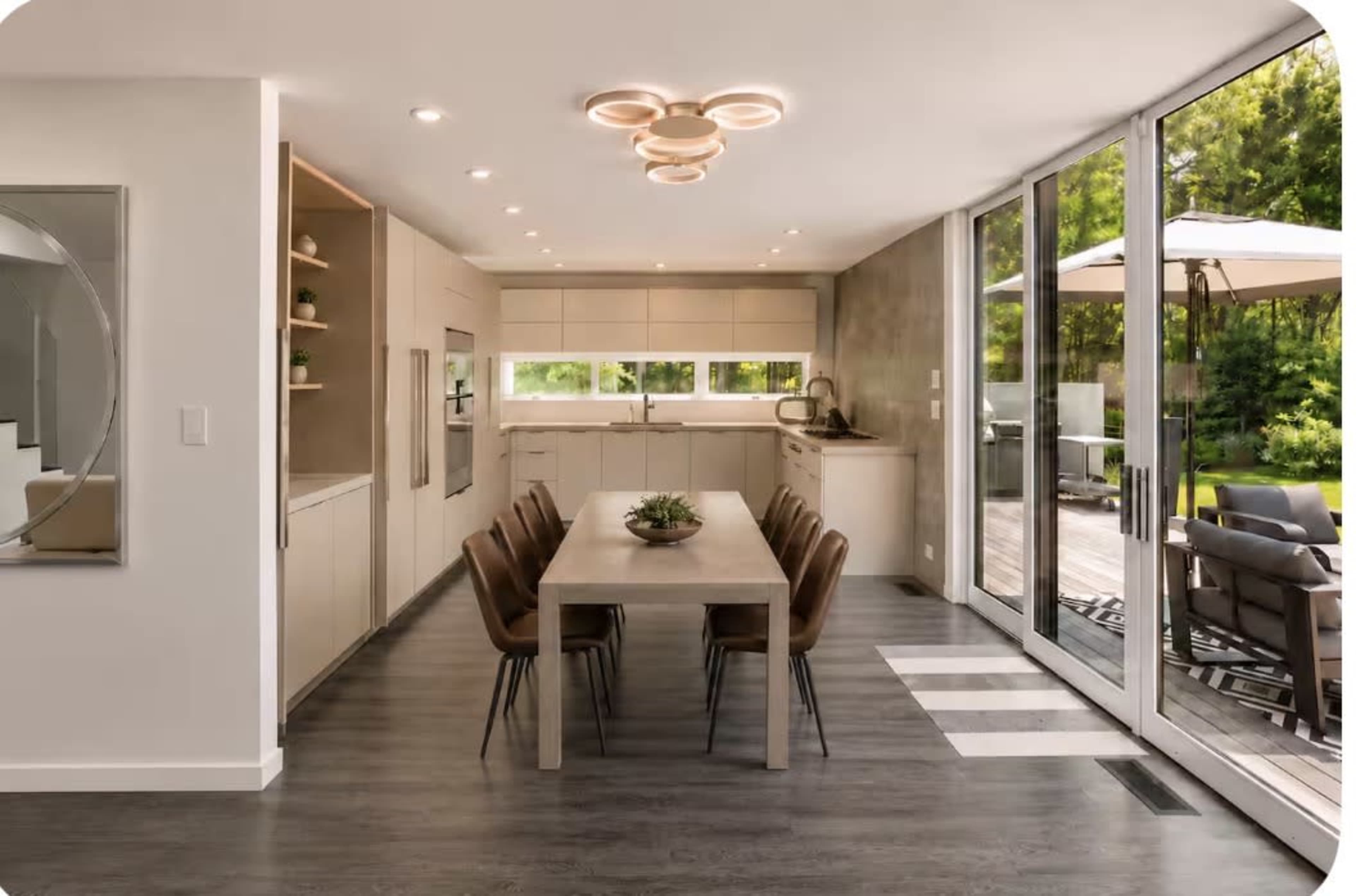 The image shows a modern kitchen and dining area featuring a sleek wooden table surrounded by chairs, with large glass doors leading to an outdoor patio.