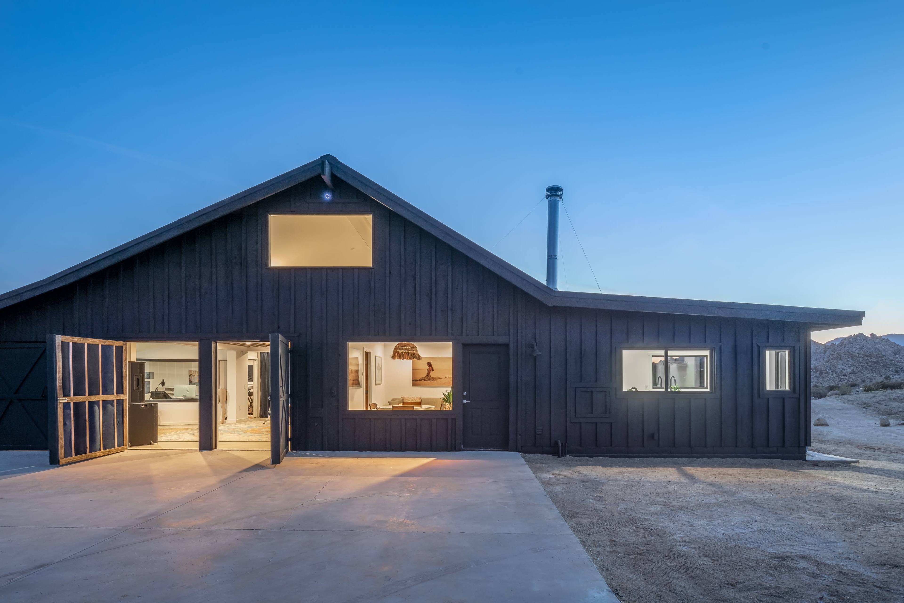 A modern black wooden house with large openings, situated in a desert landscape during twilight.