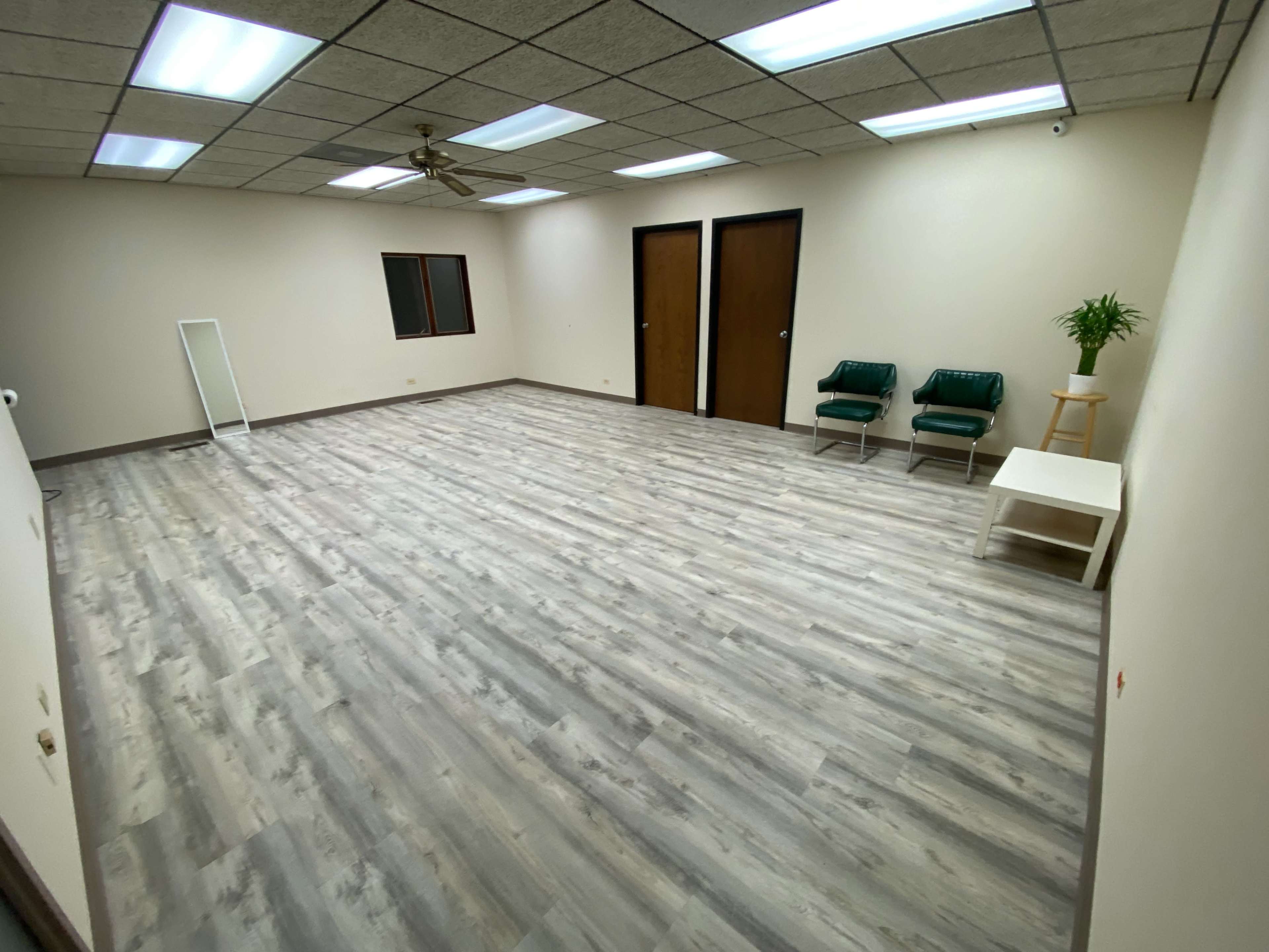 A spacious, empty room with light-colored wooden flooring, two green chairs, a small white table, and a mirror against the wall.
