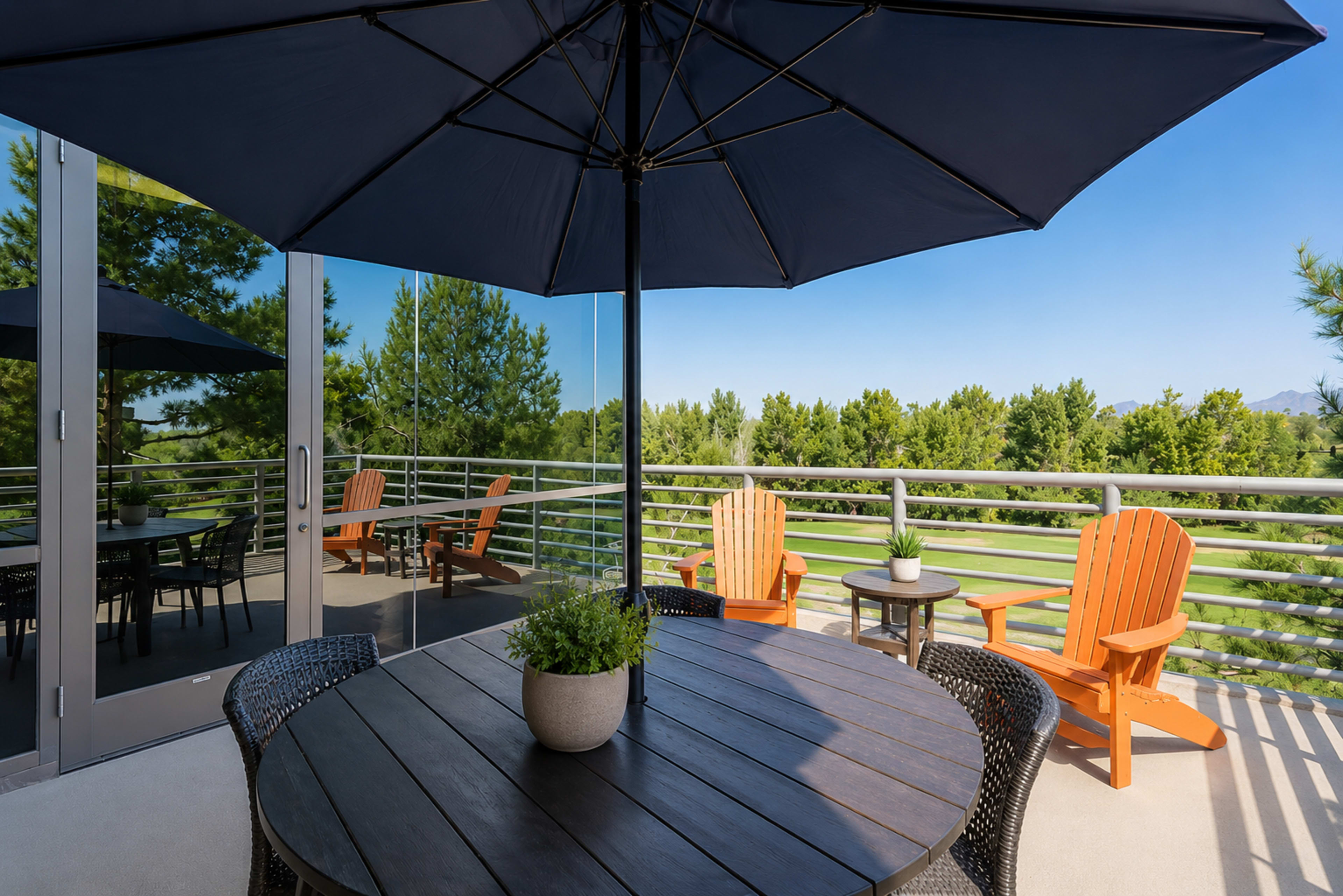 A spacious balcony features a wooden table surrounded by wicker chairs, an umbrella overhead, and orange Adirondack chairs positioned near a railing overlooking greenery.