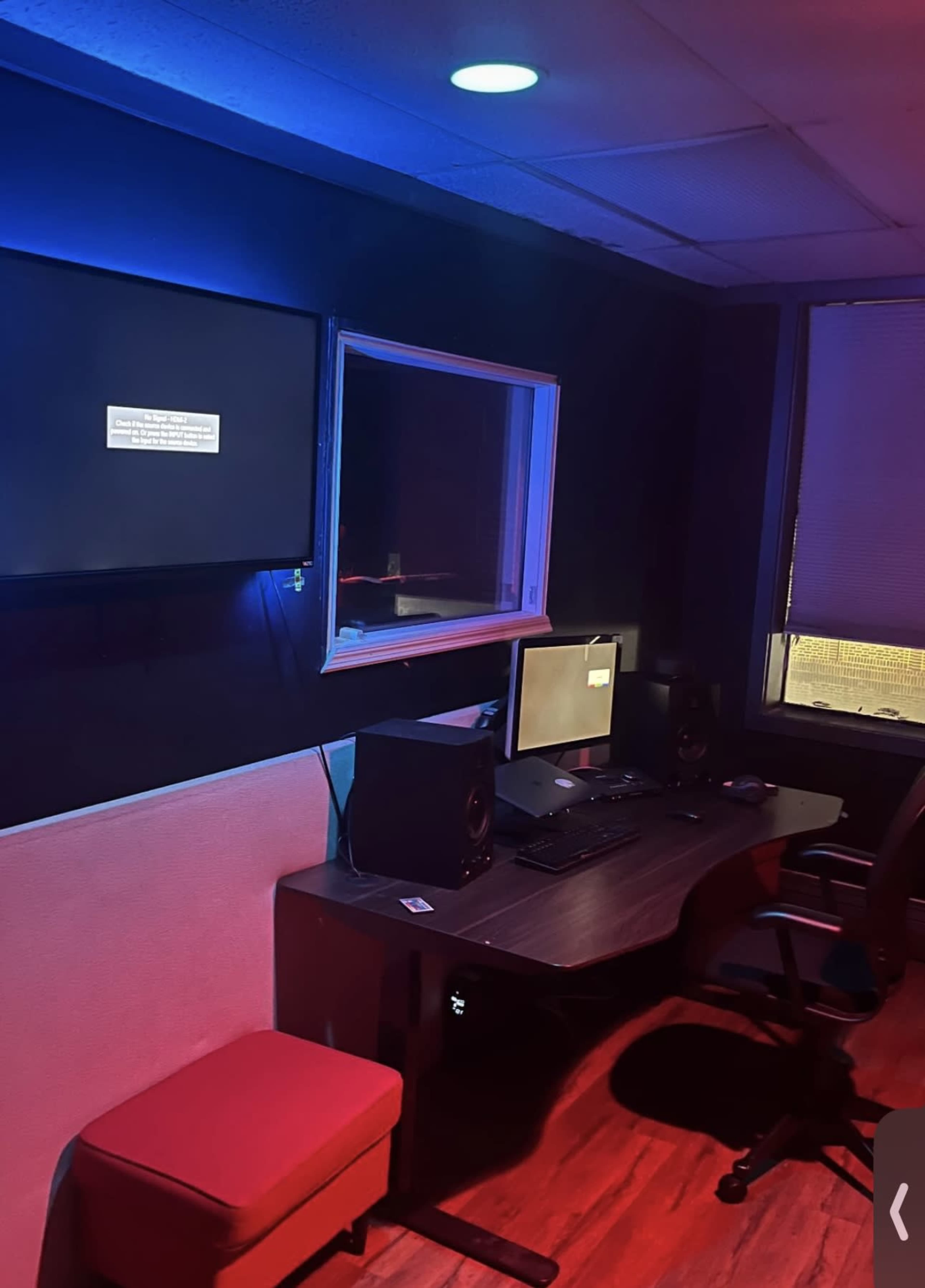 A dimly lit room contains a desk with two computer monitors, speakers, and a red chair, along with a wall-mounted screen displaying a message.