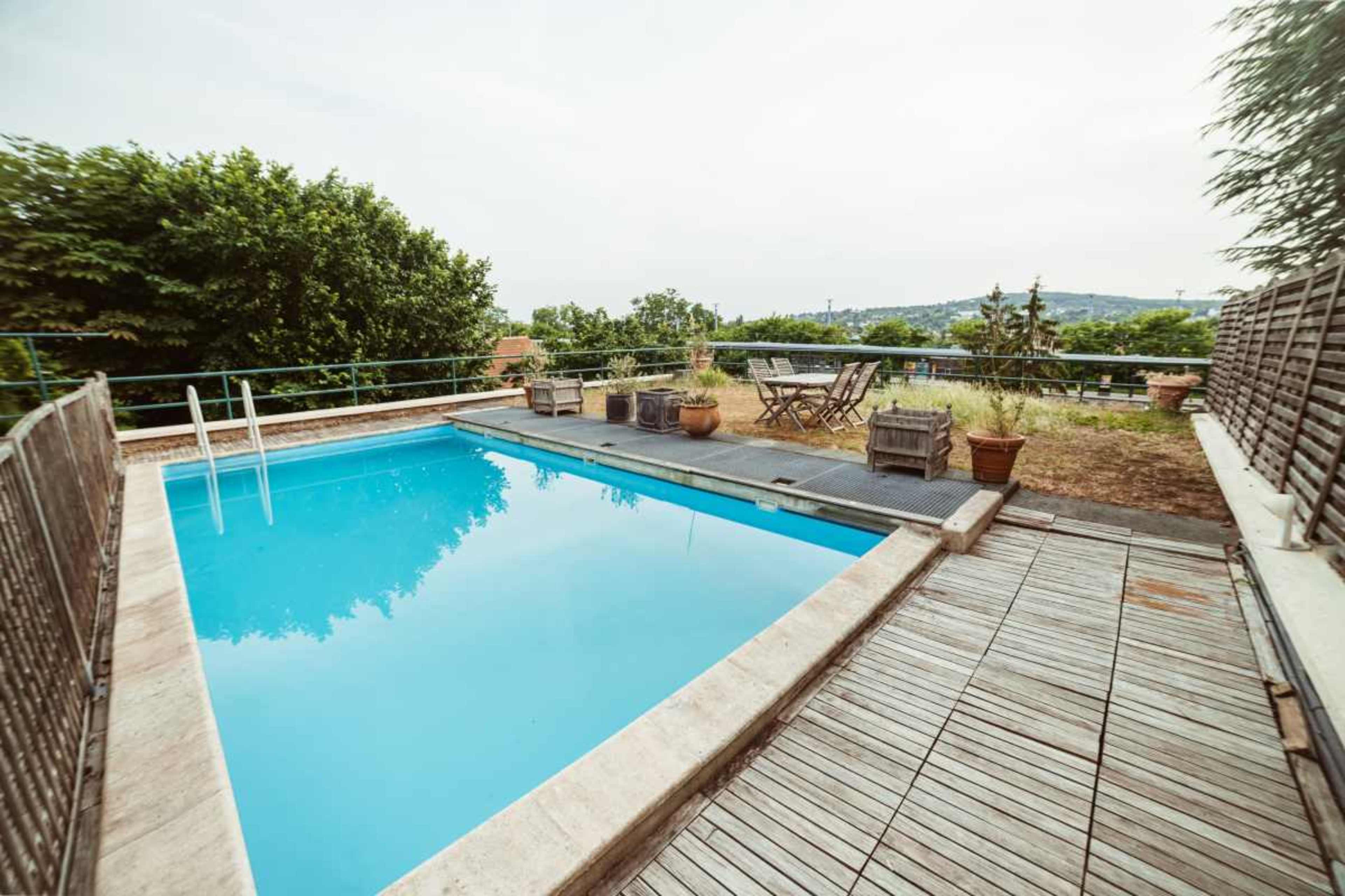 The image shows a rectangular swimming pool surrounded by wooden decking and potted plants, with a view of greenery in the background.