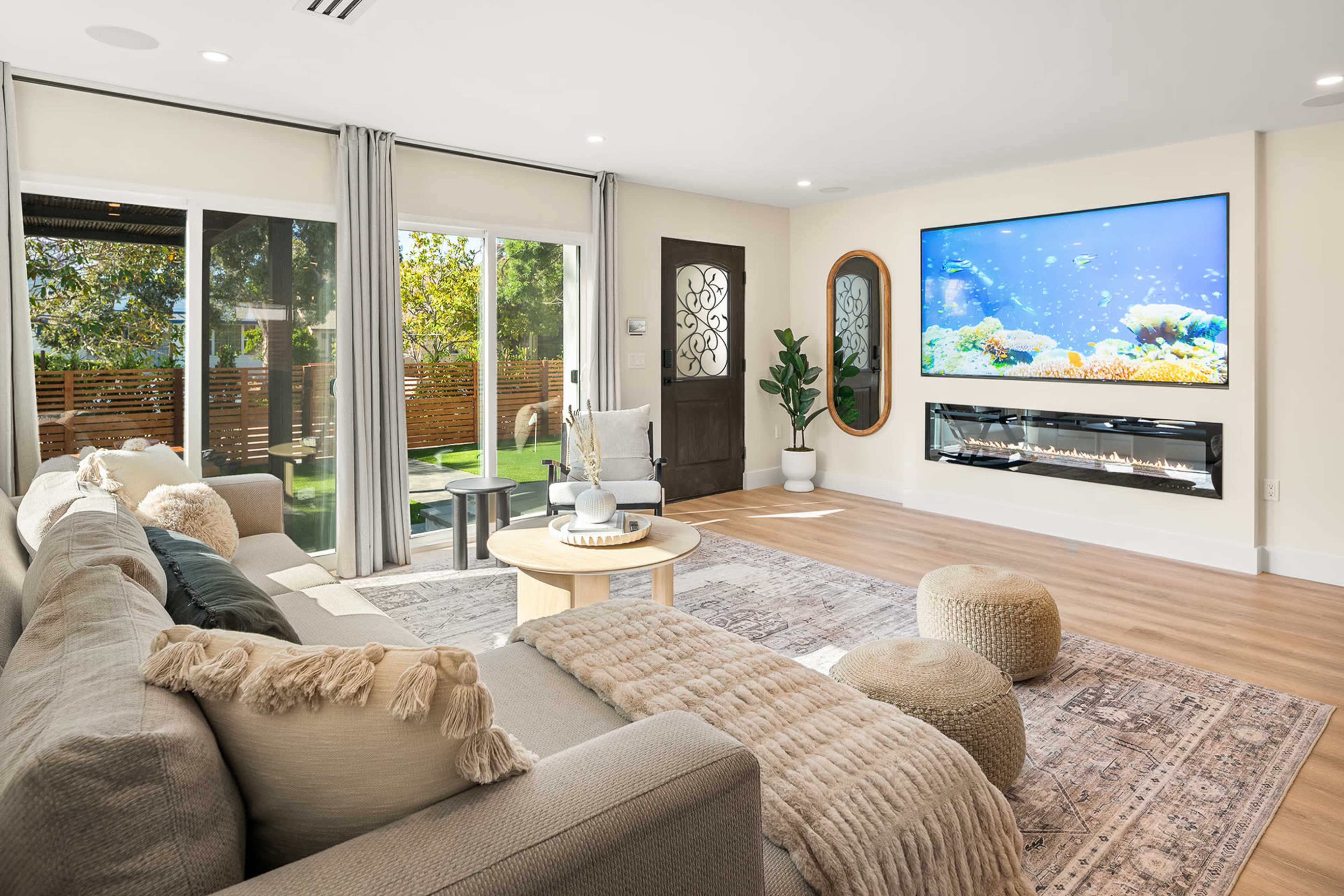 The image shows a modern living room with a large sectional sofa, a round coffee table, decorative poufs, and a wall-mounted aquarium display opposite a patio door.