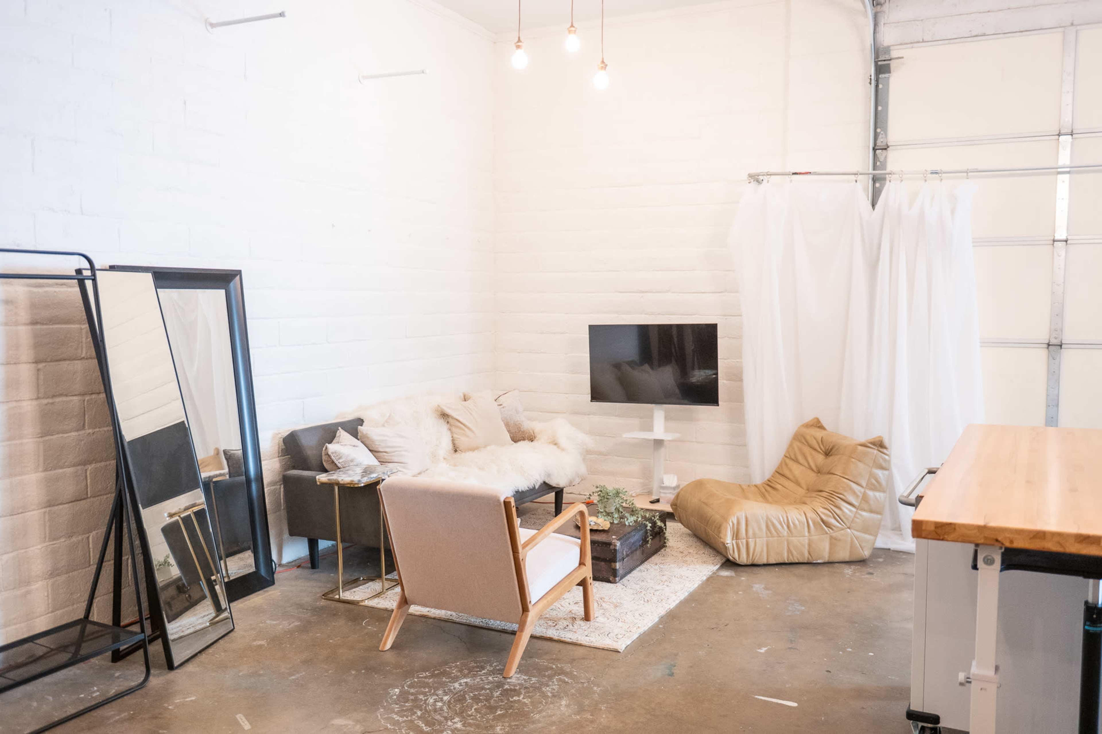 The image shows a modern, airy interior space with a cozy seating area, including a sofa with throw pillows, a chair, and a large bean bag, along with a wall-mounted television and a large mirror.