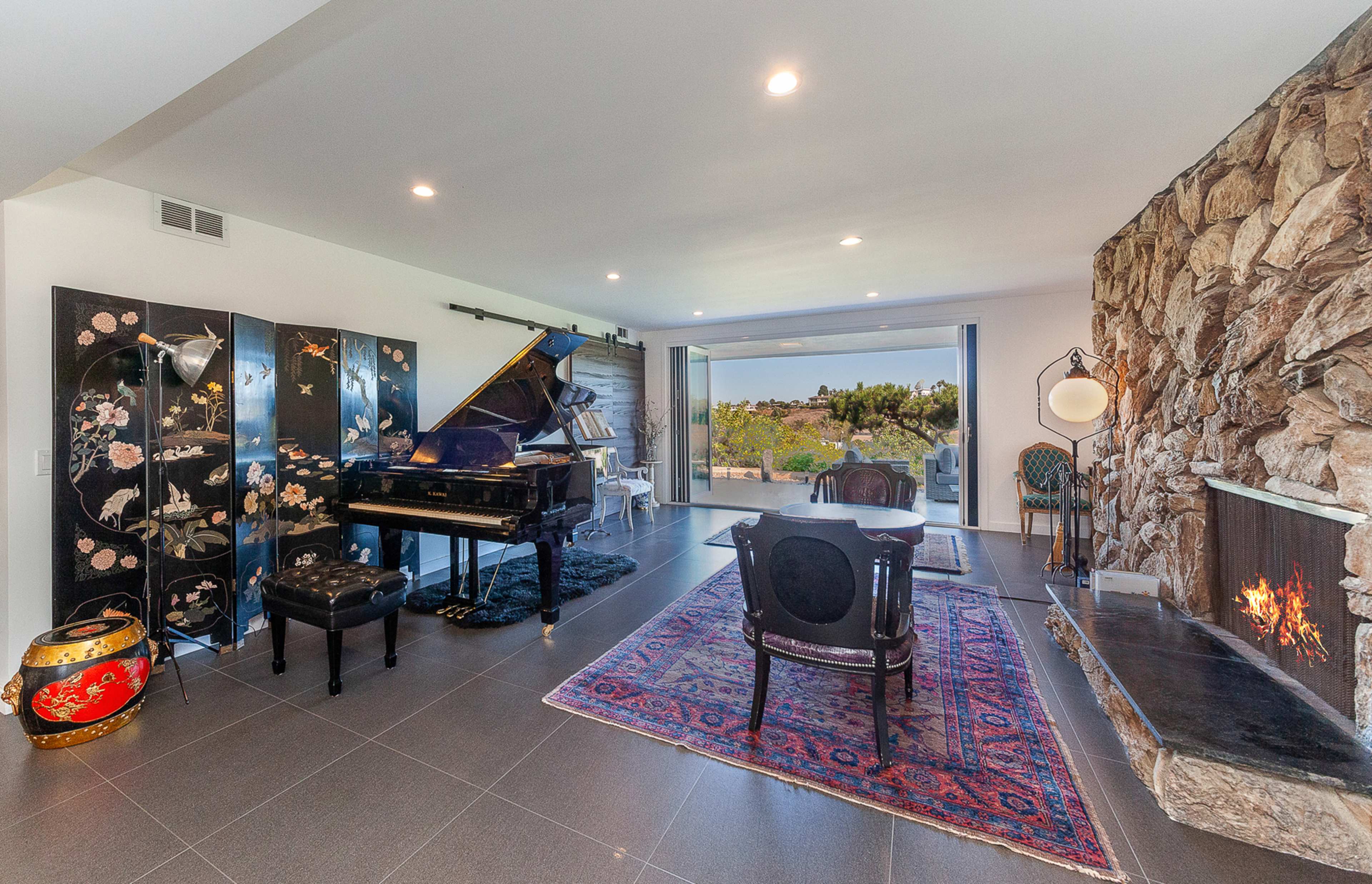 The room features a grand piano beside a stone fireplace, with a black screen adorned with floral designs and large sliding glass doors opening to a view of the outdoors.
