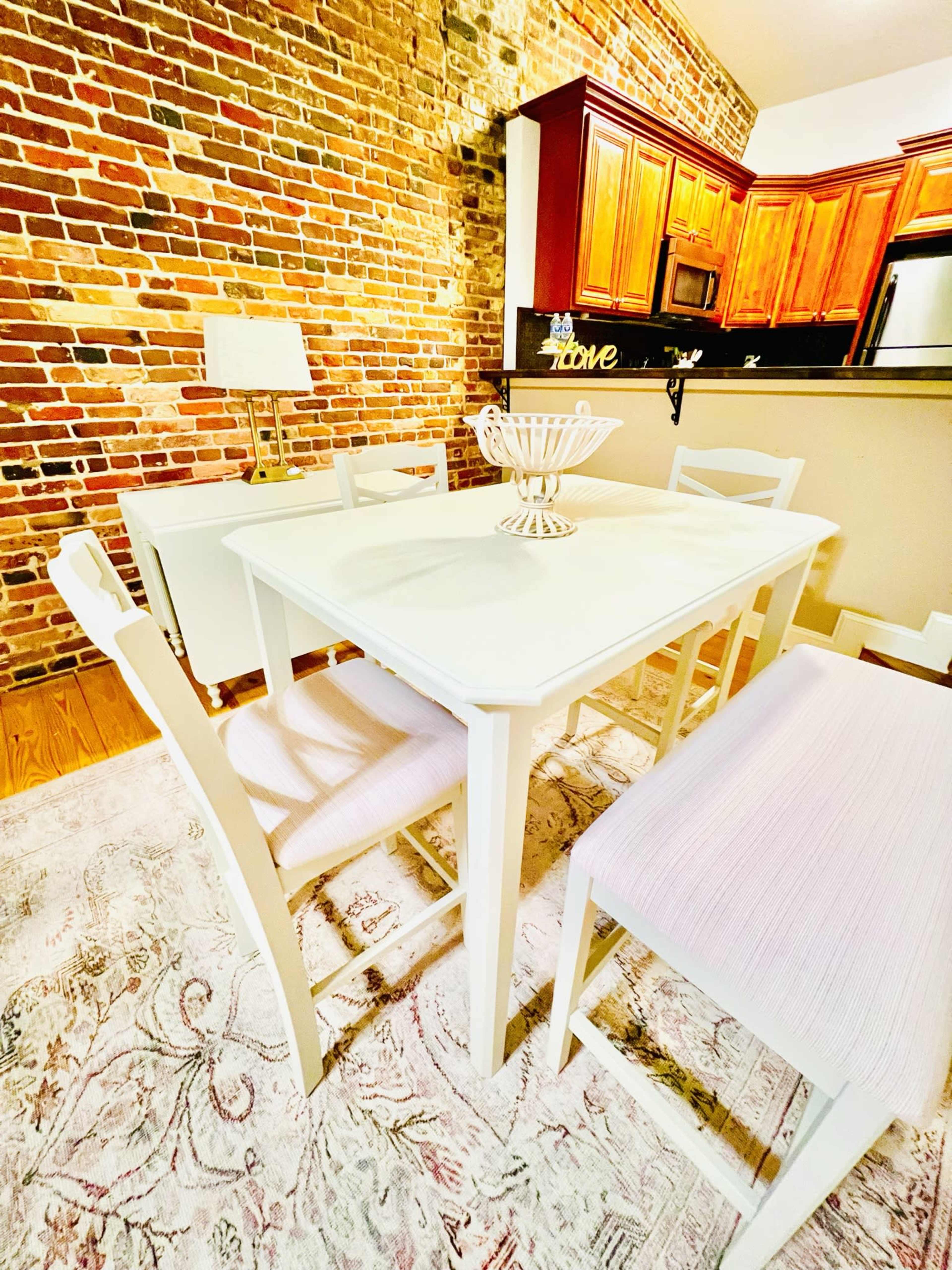 A white dining table surrounded by wooden chairs on a patterned rug, with a brick wall and kitchen cabinetry in the background.