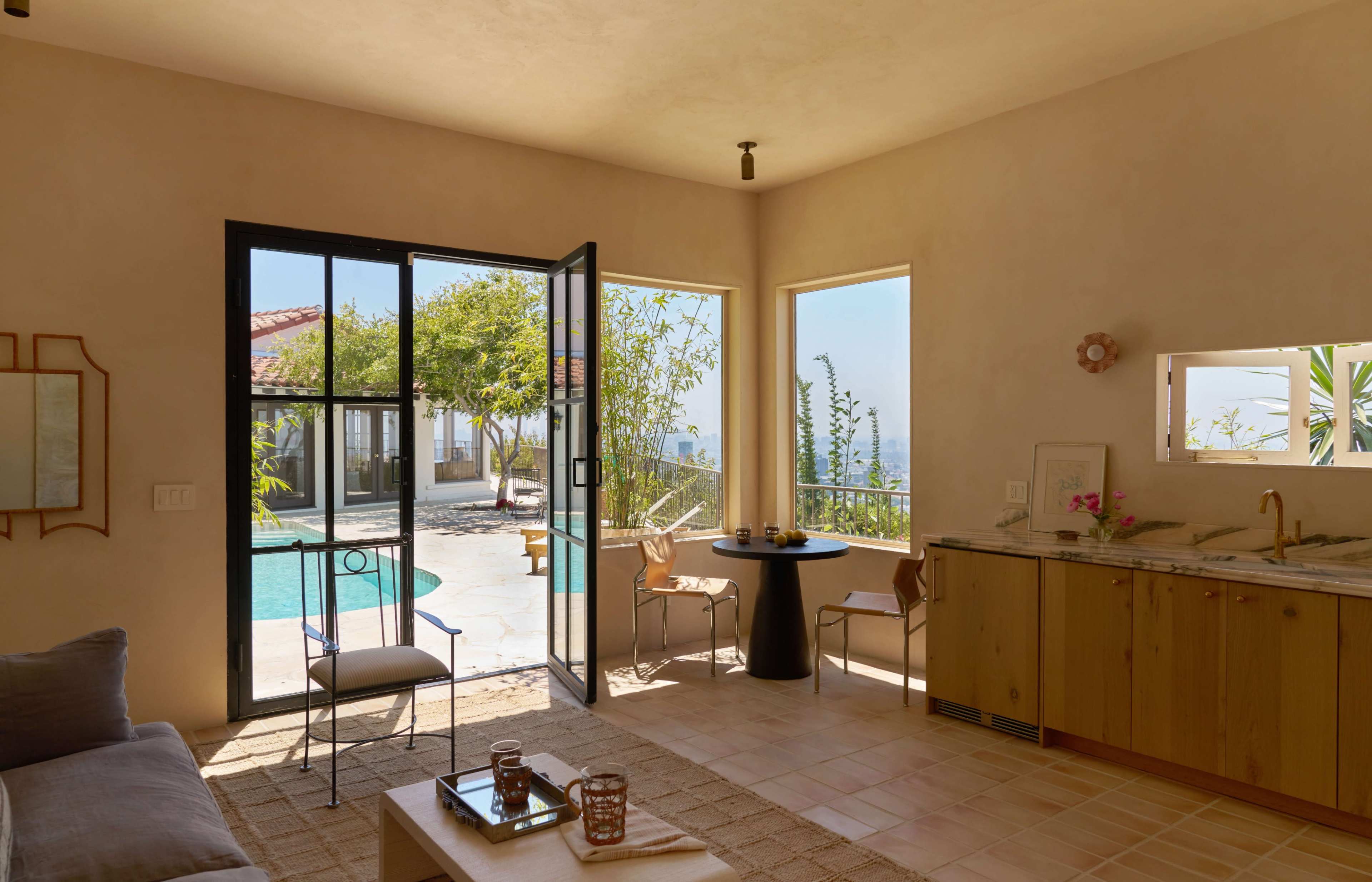 A bright indoor space features a kitchen area, a small dining table, and large open doors leading to a backyard pool surrounded by greenery.