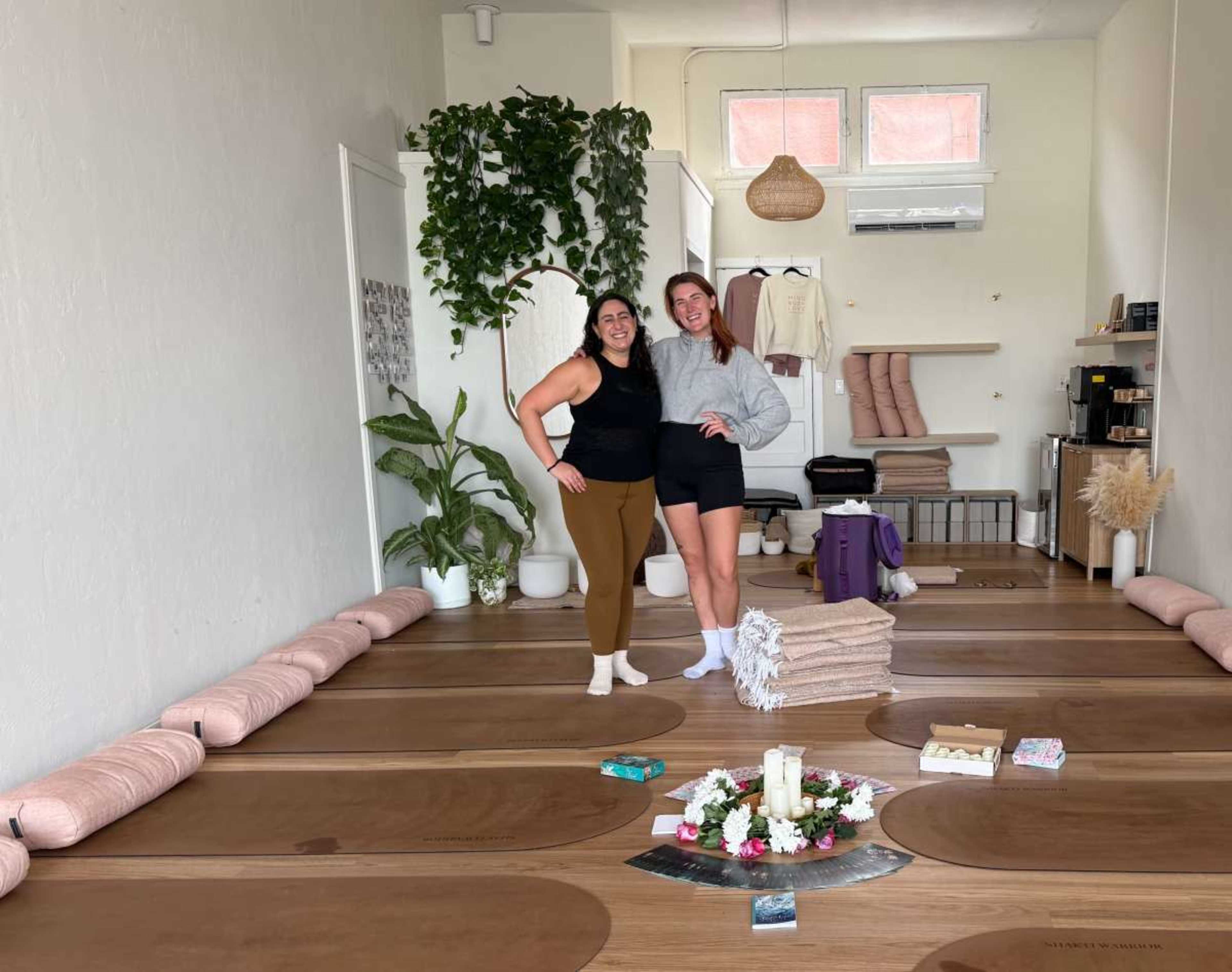 Two women pose together in a yoga studio, surrounded by yoga mats, cushions, and decorative plants.