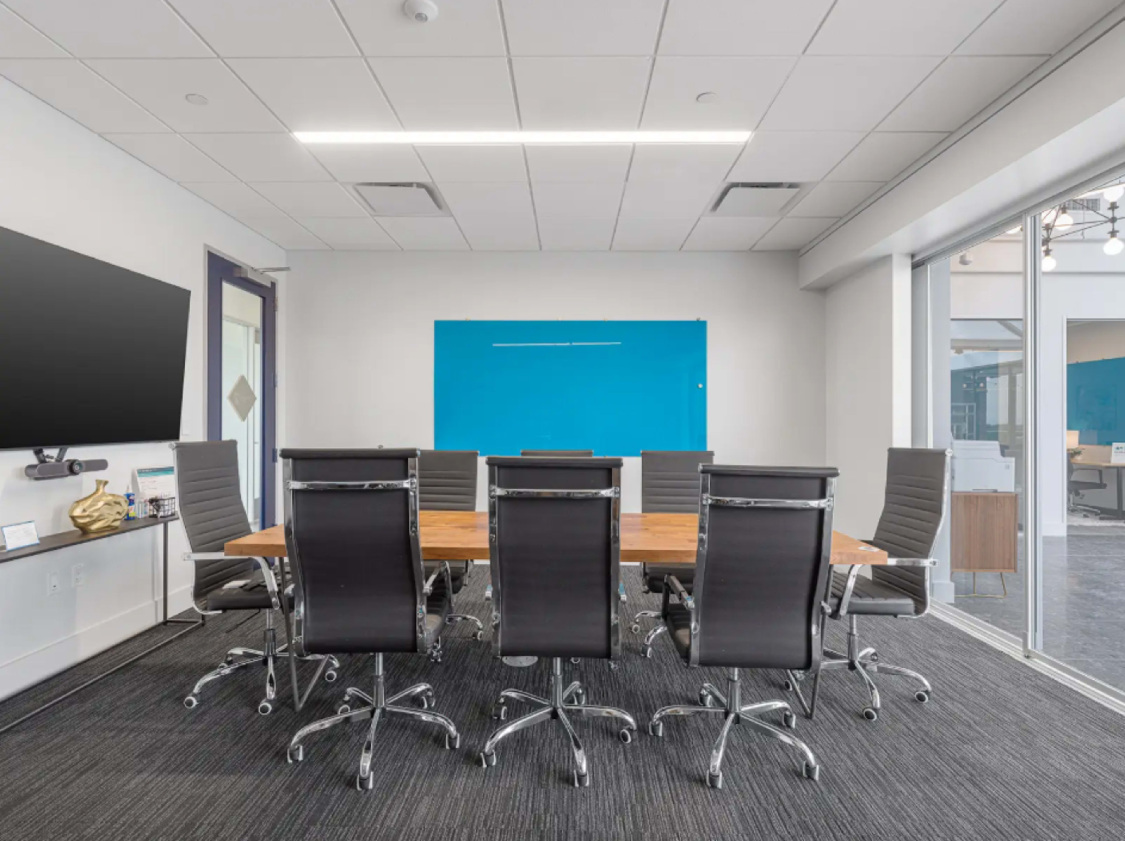The image shows a modern conference room with a long wooden table surrounded by black wheeled chairs and a blue accent wall.