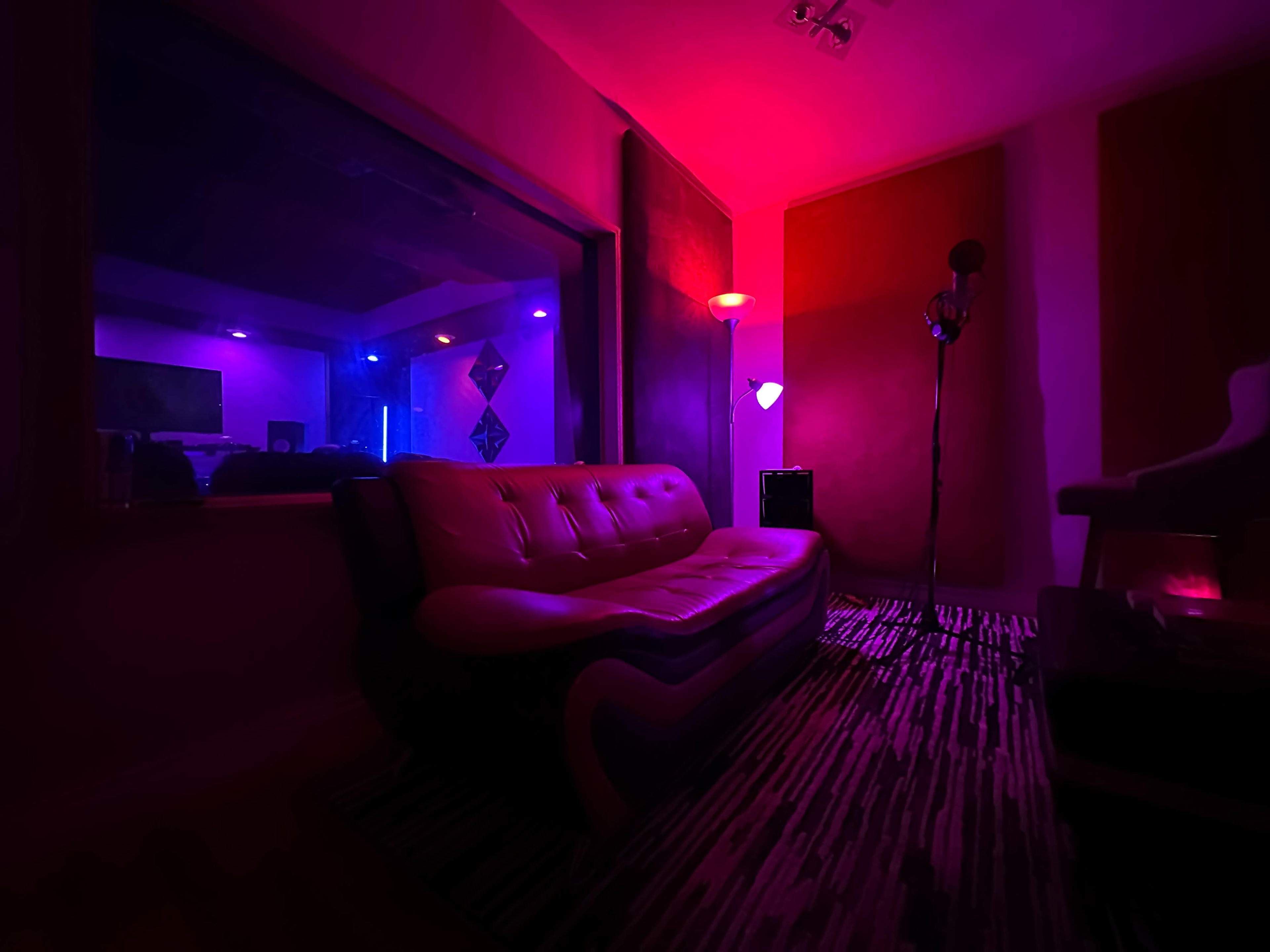 A dimly lit music studio with a red and purple color scheme, featuring a modern couch, a microphone stand, and various studio equipment visible through a glass window.