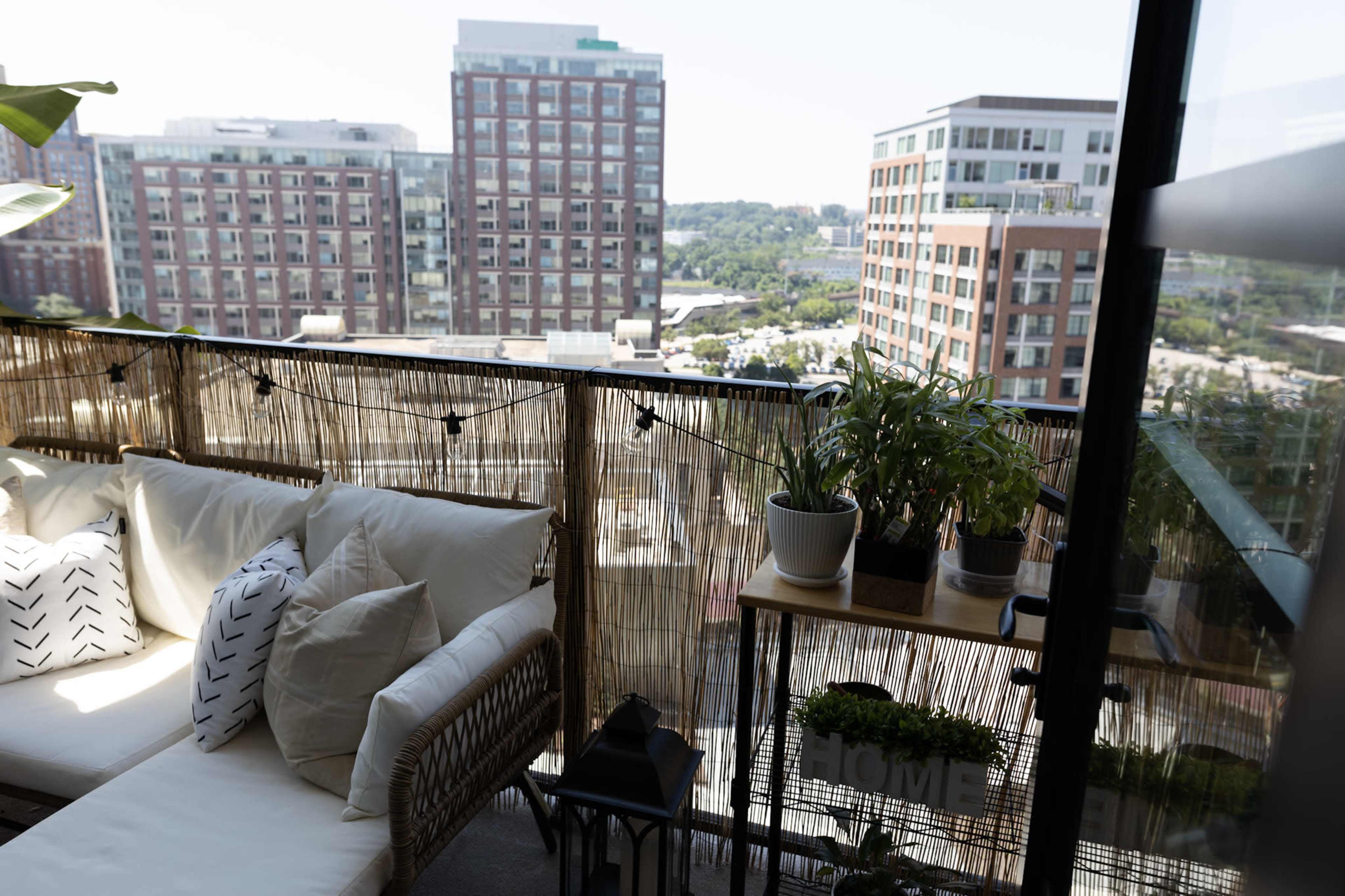 A balcony features a wicker sofa adorned with pillows, potted plants, and a view of surrounding buildings and greenery.