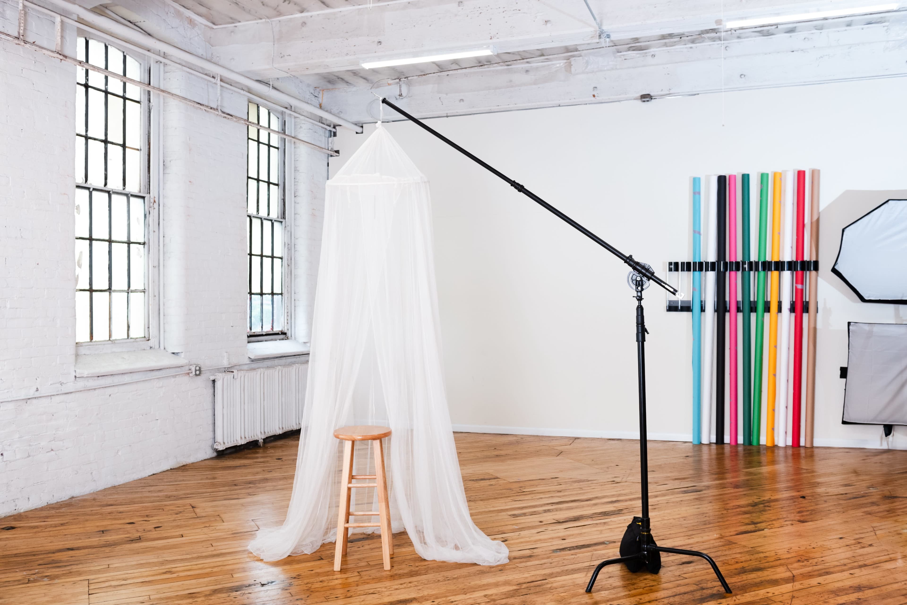 A well-lit studio space featuring a stool draped with a sheer white fabric suspended from a lighting stand, alongside colorful backdrops mounted on the wall.