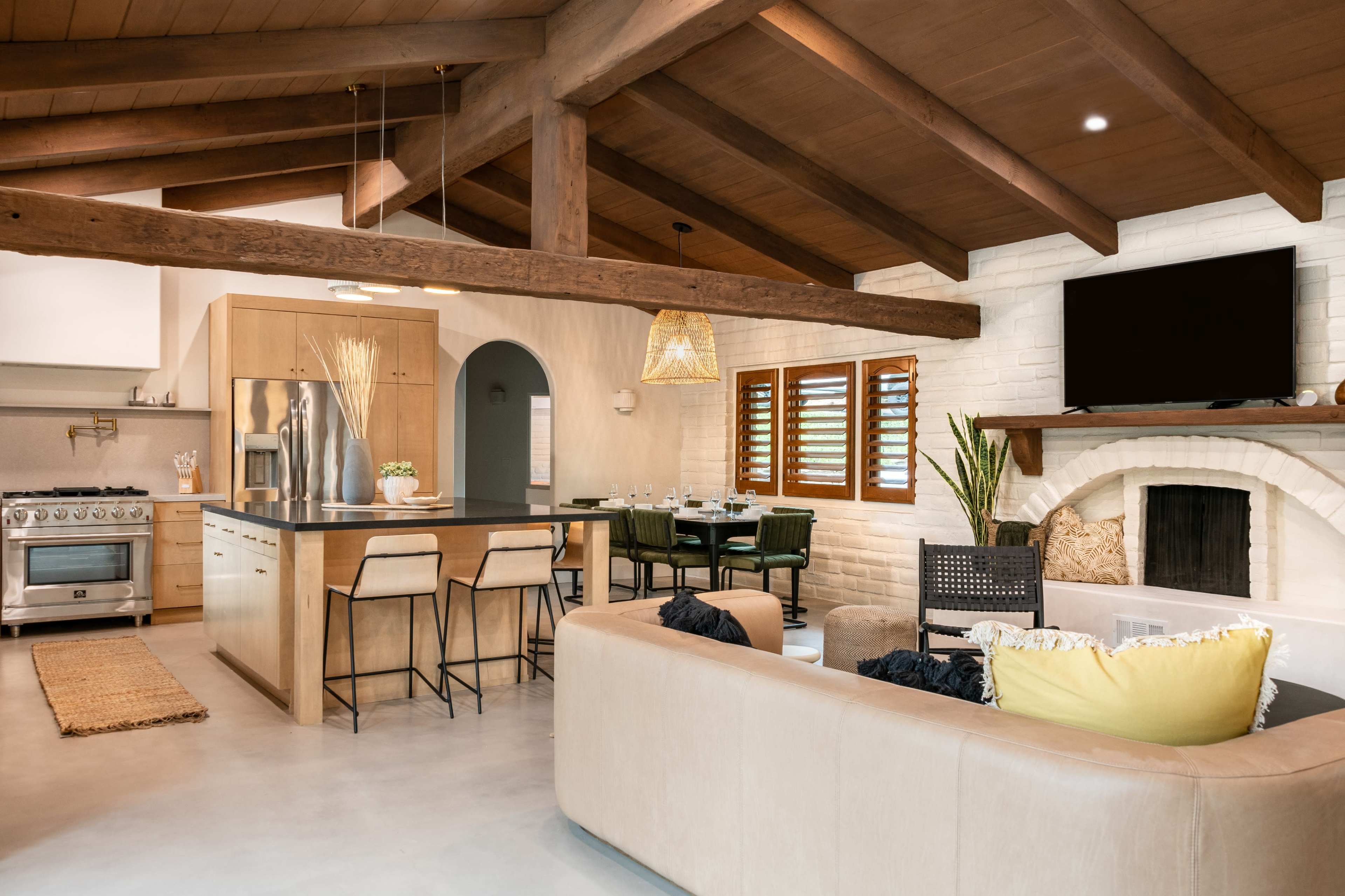 A modern kitchen and living area featuring wooden beams, a large island with bar stools, and a cozy seating arrangement with a television mounted on the wall.