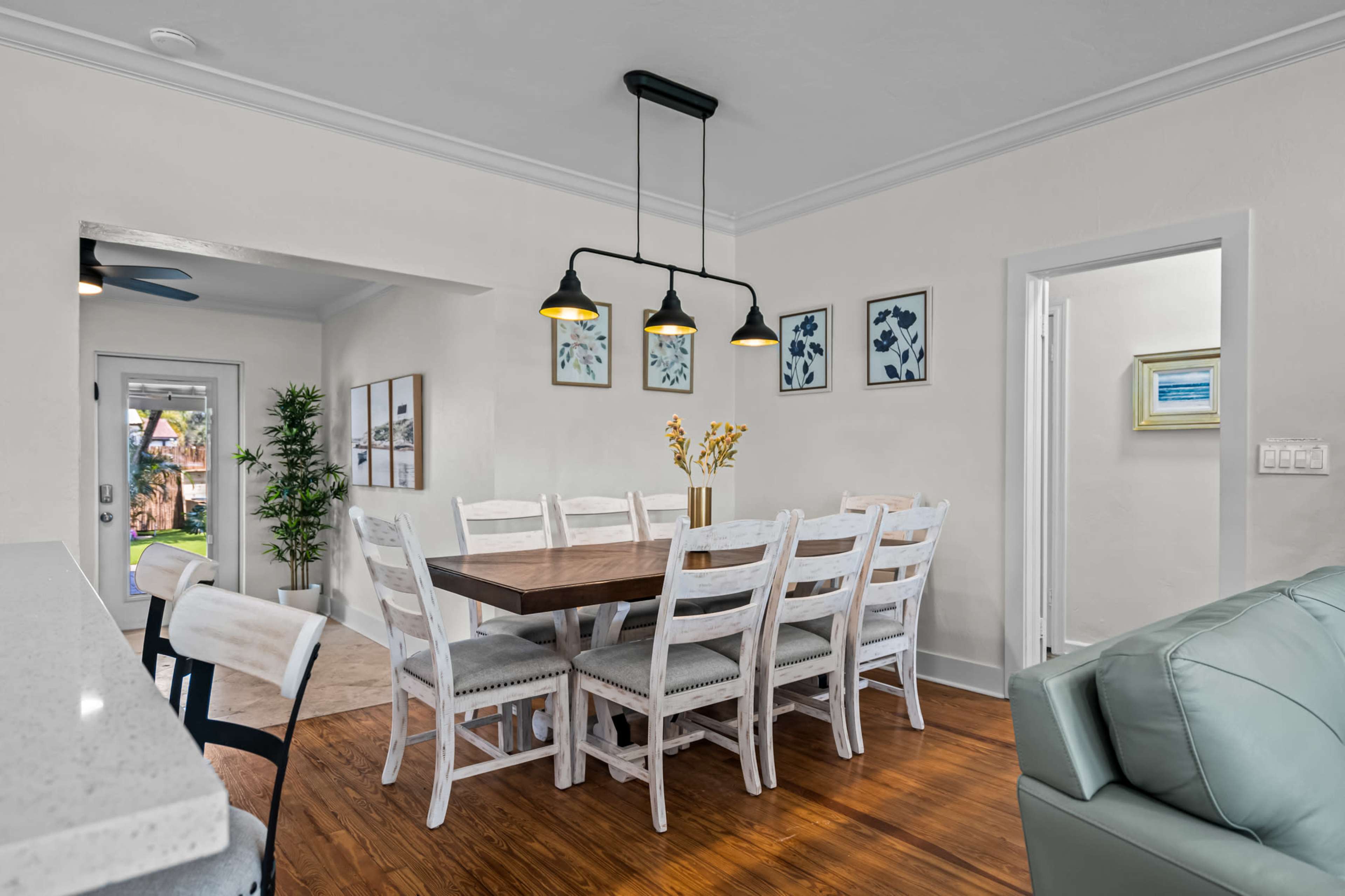 A dining area features a large wooden table surrounded by white chairs, illuminated by pendant lights, with framed artwork on the walls.
