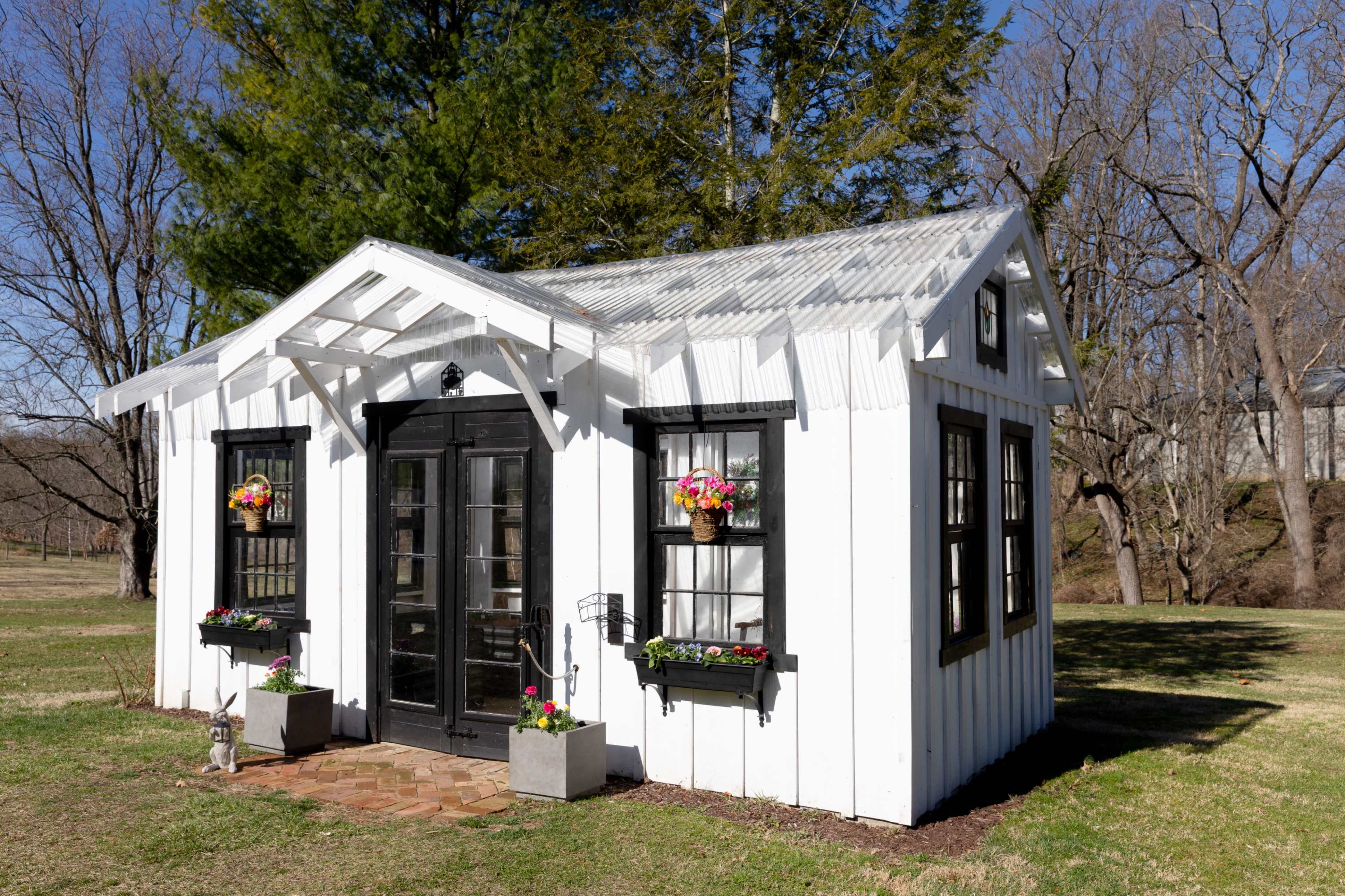 A small, white garden shed with a gabled roof and decorative flower boxes is situated in a grassy area surrounded by bare trees.