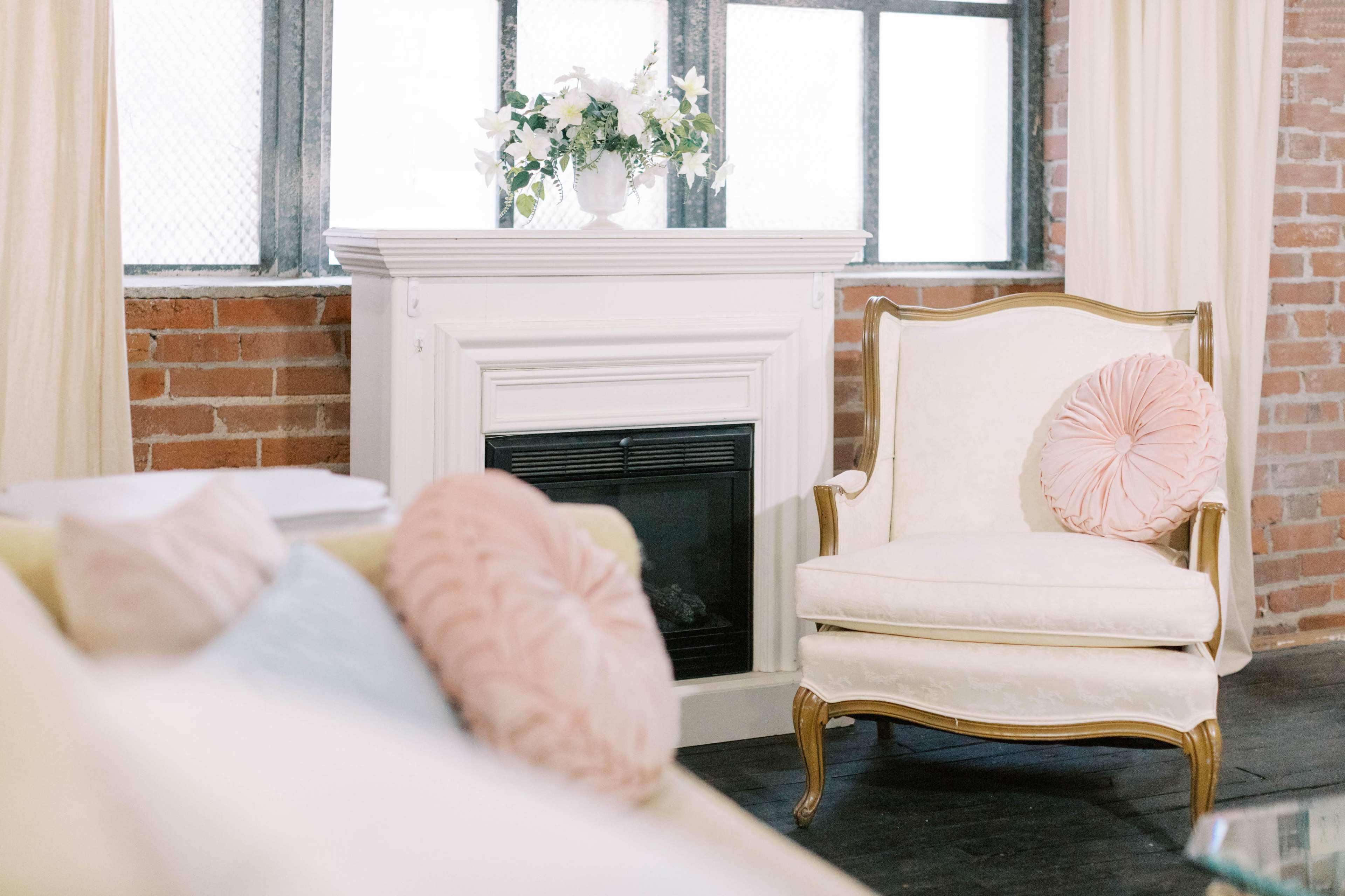 A cozy seating area featuring a white armchair with a pink decorative pillow beside a white fireplace and a window adorned with light curtains.