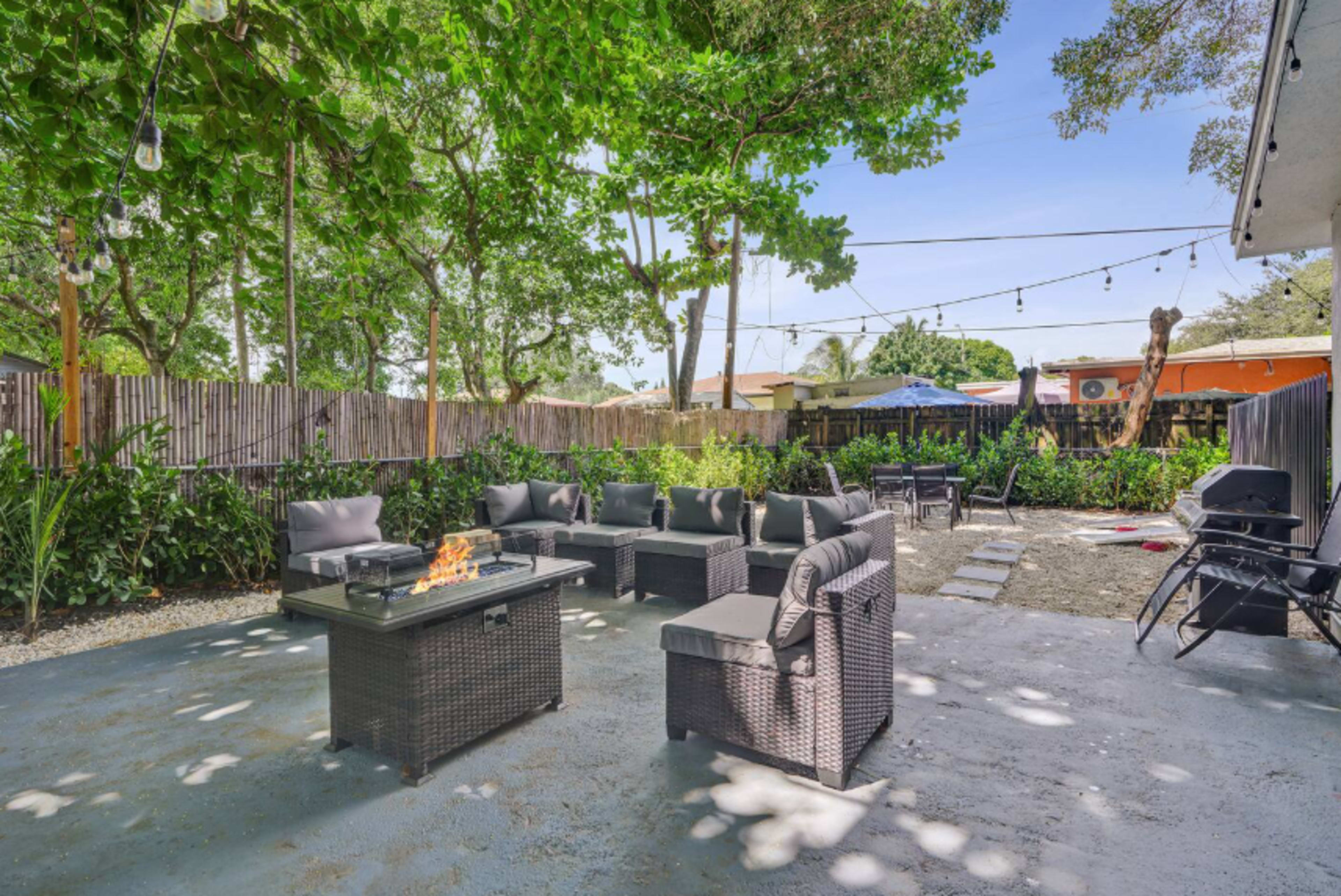 The image shows an outdoor seating area with dark wicker furniture arranged around a fire pit, surrounded by trees and a fence, with string lights overhead.