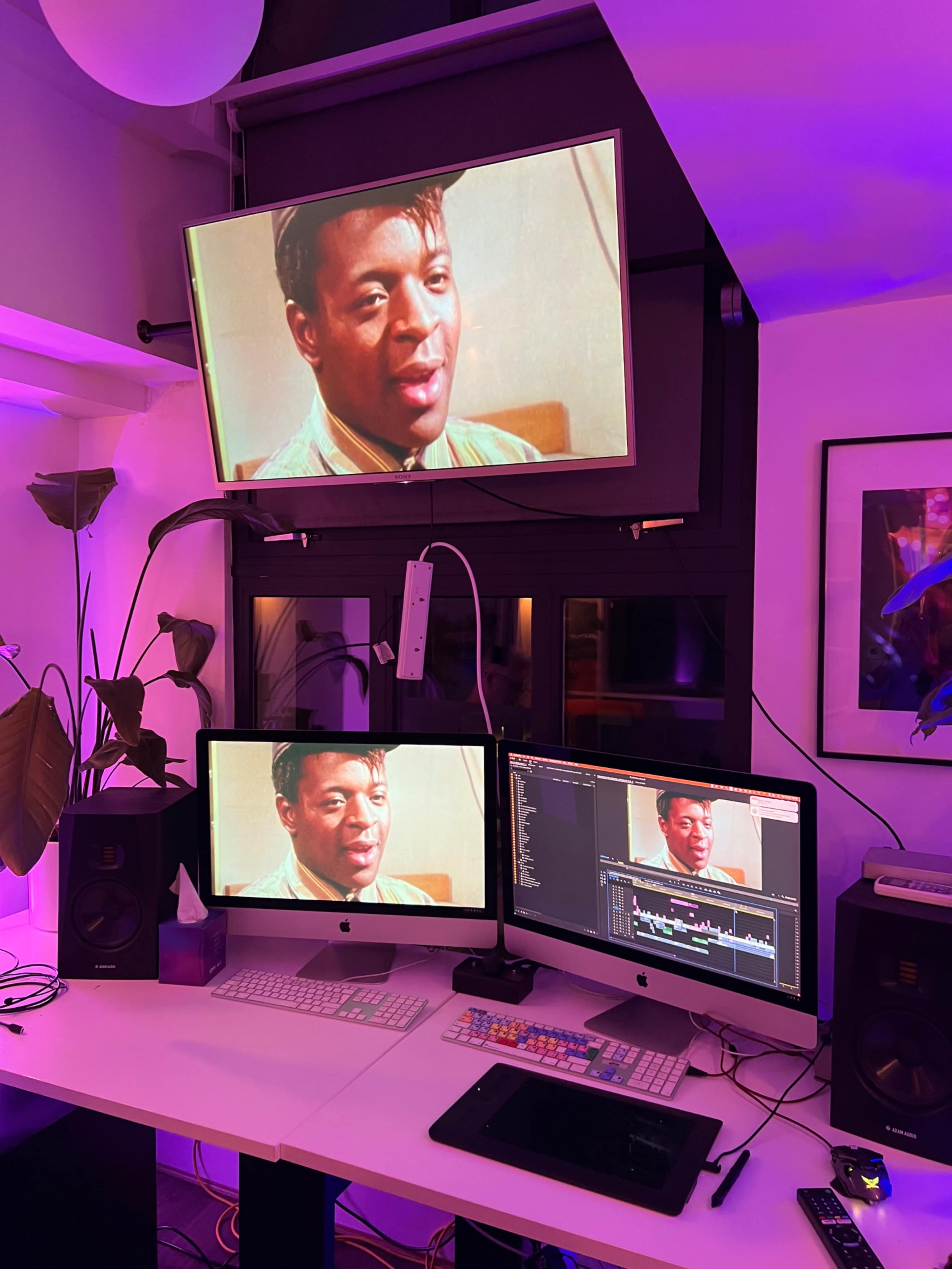 The image shows a modern workspace with three screens displaying a man's face, illuminated by purple lighting and featuring plants in the background.