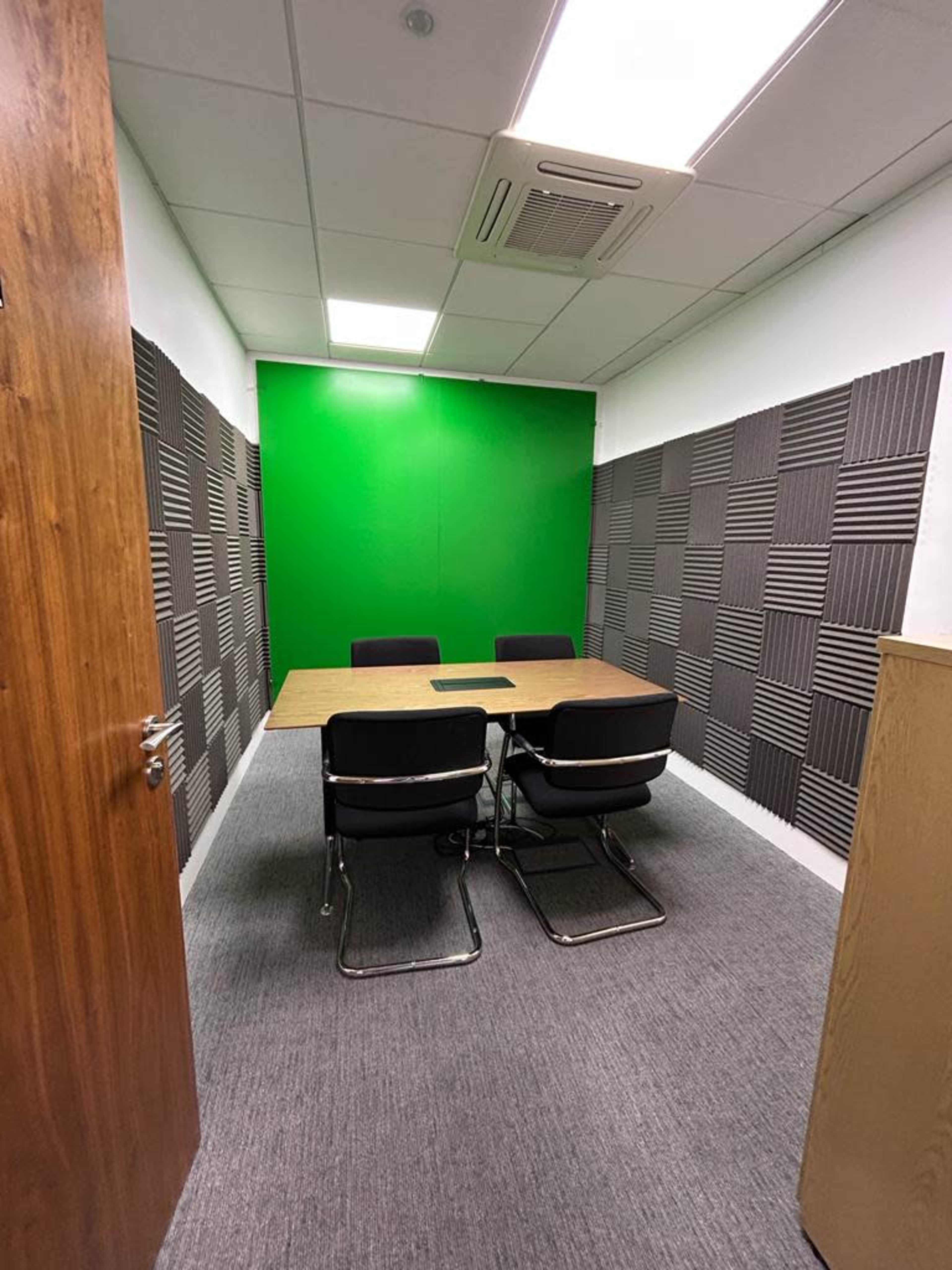 A small meeting room with a wooden table and four black chairs, featuring soundproof panels on the walls and a bright green backdrop.
