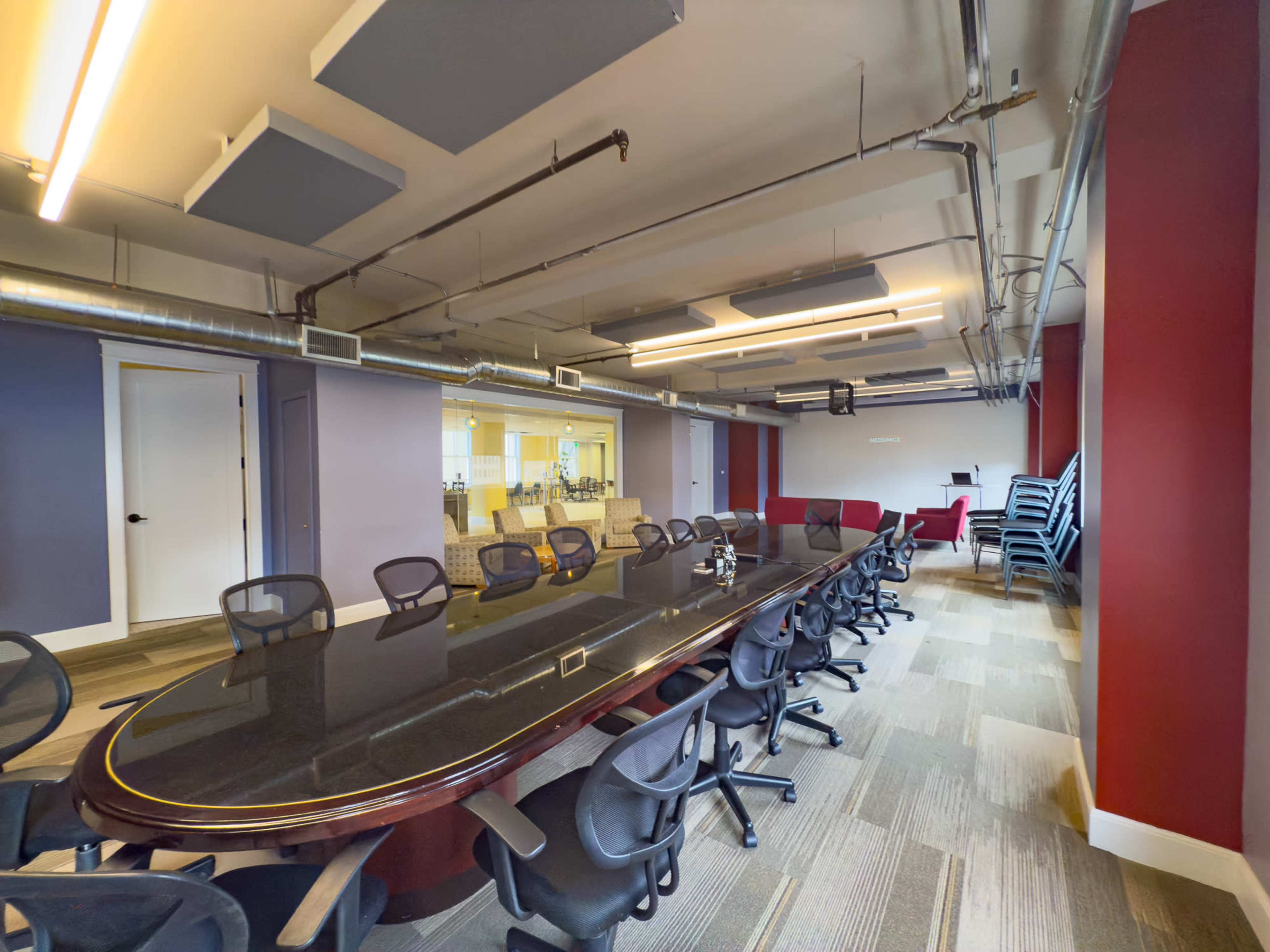 The image shows a modern conference room featuring a large black table surrounded by office chairs, with walls in varying colors and stacked chairs in the background.