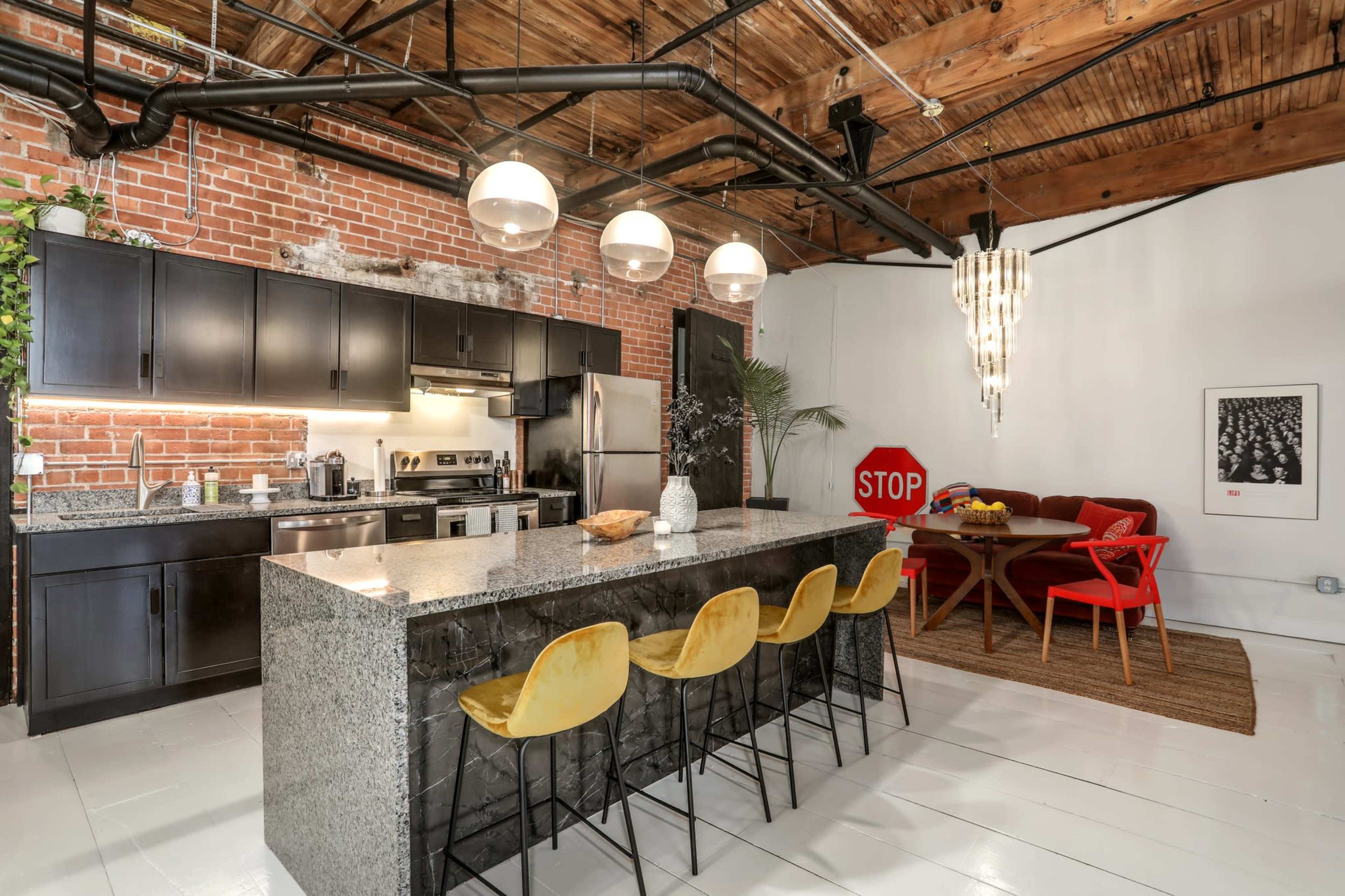 A modern kitchen with a granite island and bar seating, featuring exposed brick walls and industrial-style lighting, alongside a dining area with a red table and chairs.