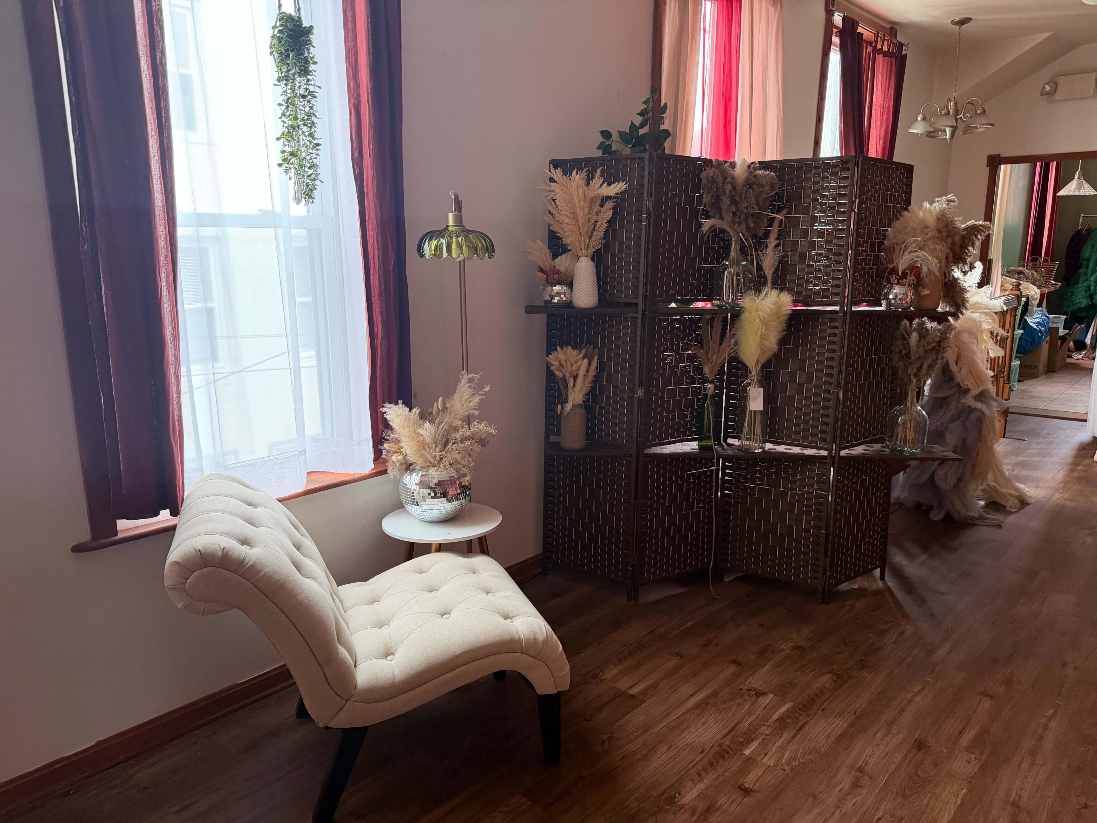 The image features a cozy room with a tufted beige chair, a small side table, a decorative room divider with dried plants, and large windows draped with red curtains.