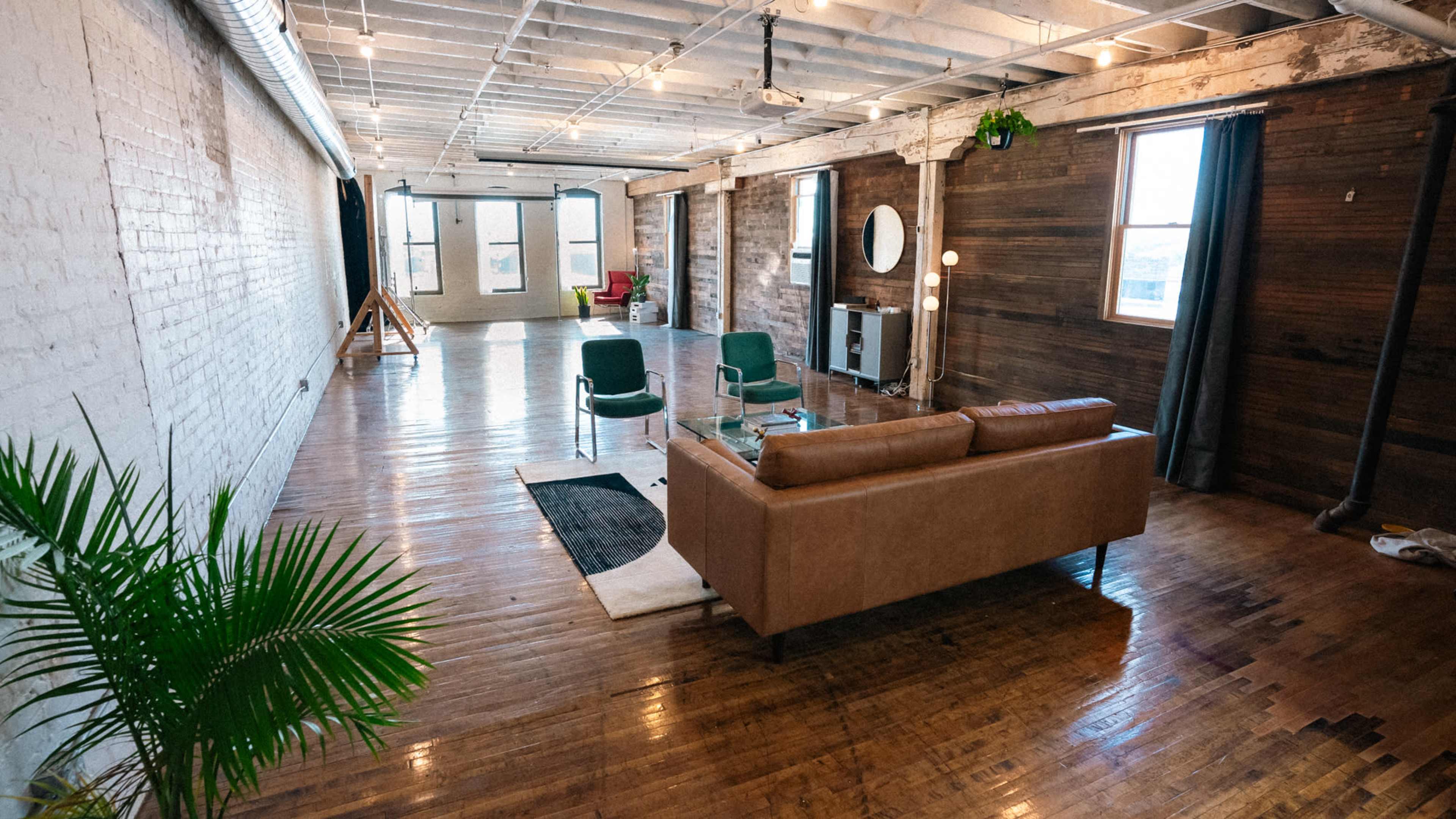A spacious interior with hardwood floors, featuring a brown sofa, green chairs, a plant, and large windows that illuminate the room.