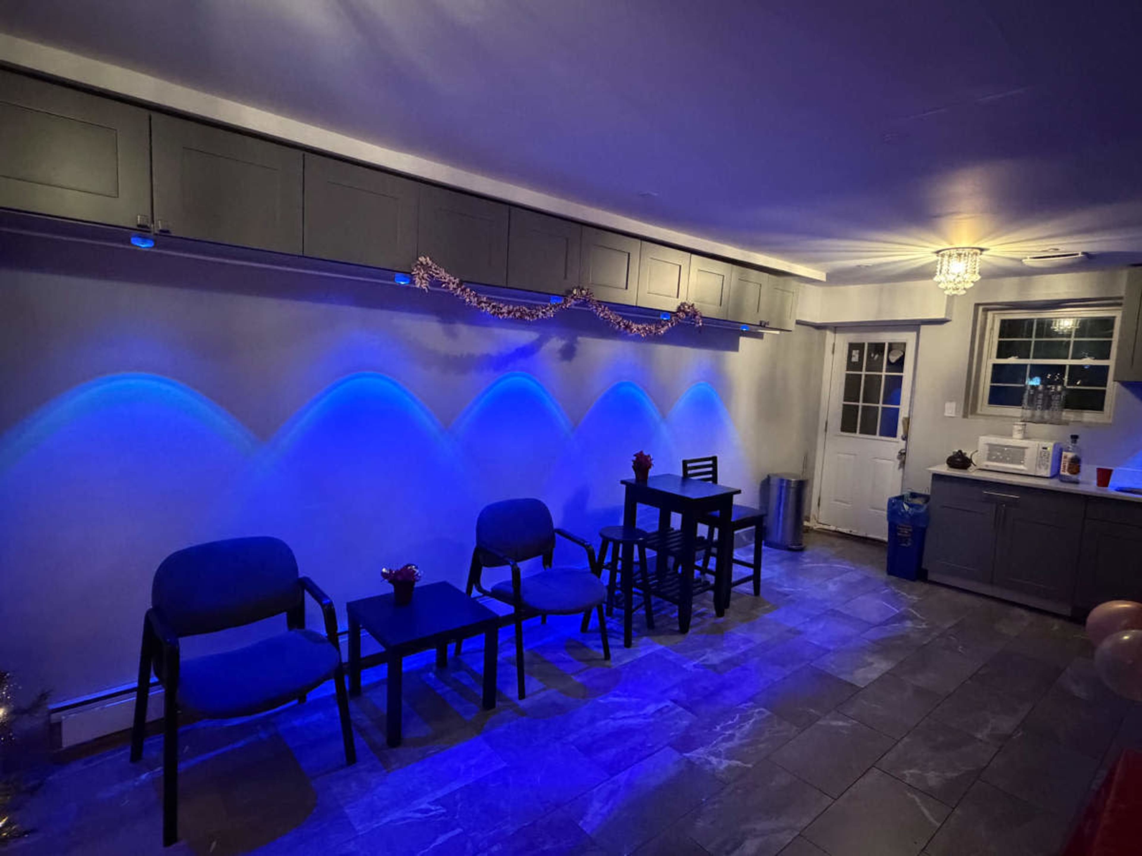 The image shows a room with gray cabinetry and blue lighting, featuring a seating area with two chairs and a small table, along with a door and a kitchenette on one side.