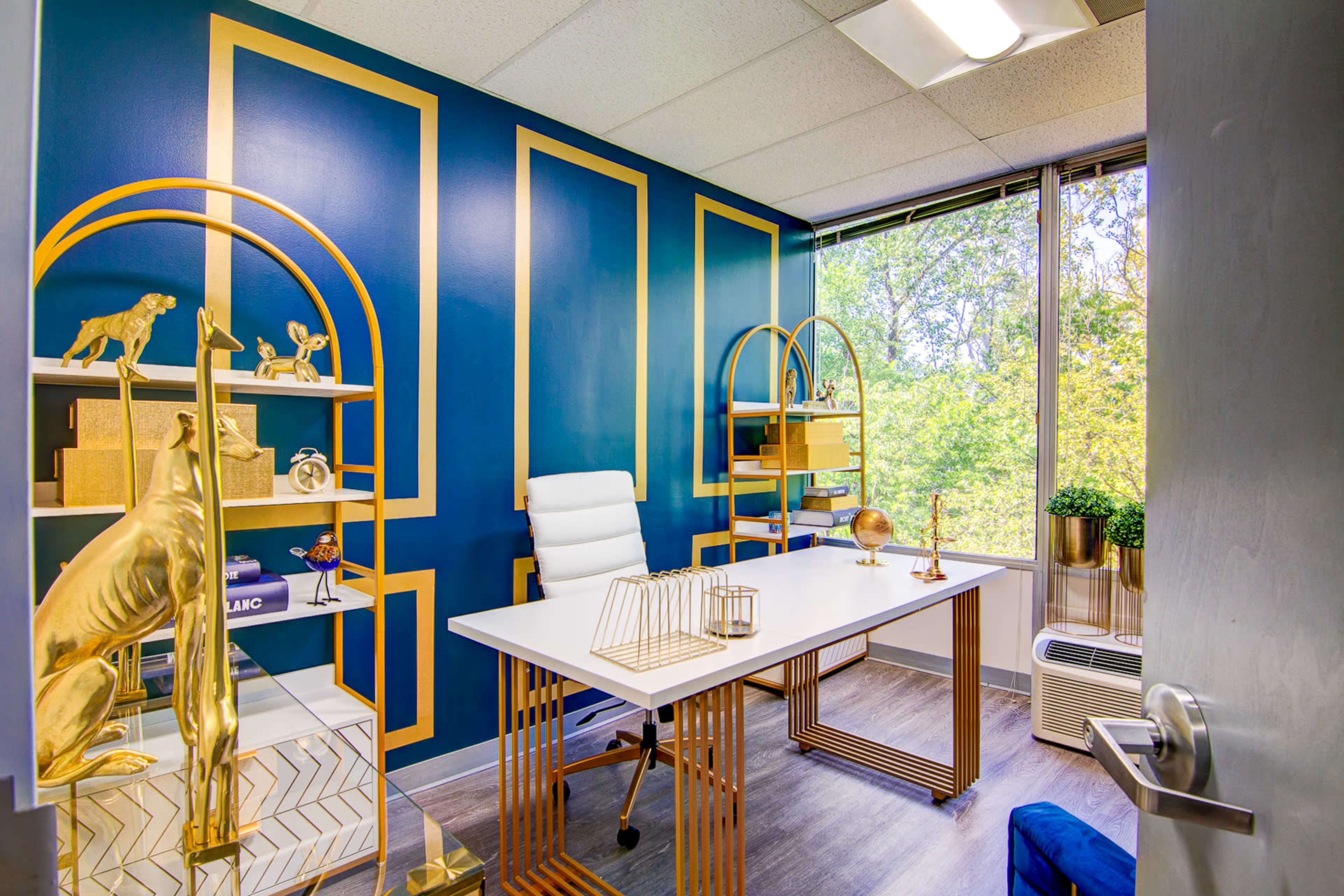 A modern office with a white desk, gold accents, and large windows overlooking greenery.