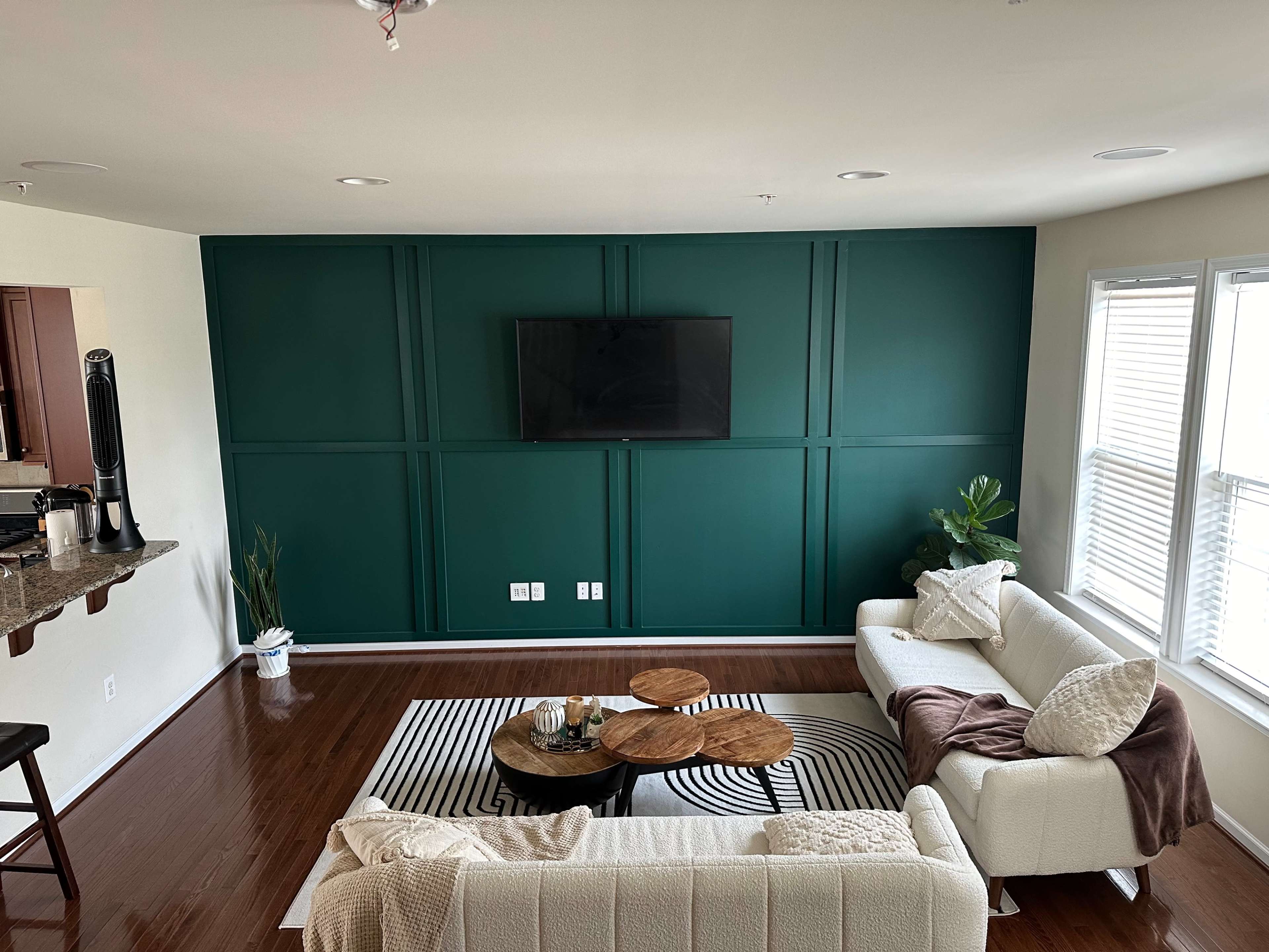 A modern living room with a green accent wall, a flat-screen TV, two cream-colored sofas, a round coffee table with wooden surfaces, and a striped black-and-white rug.