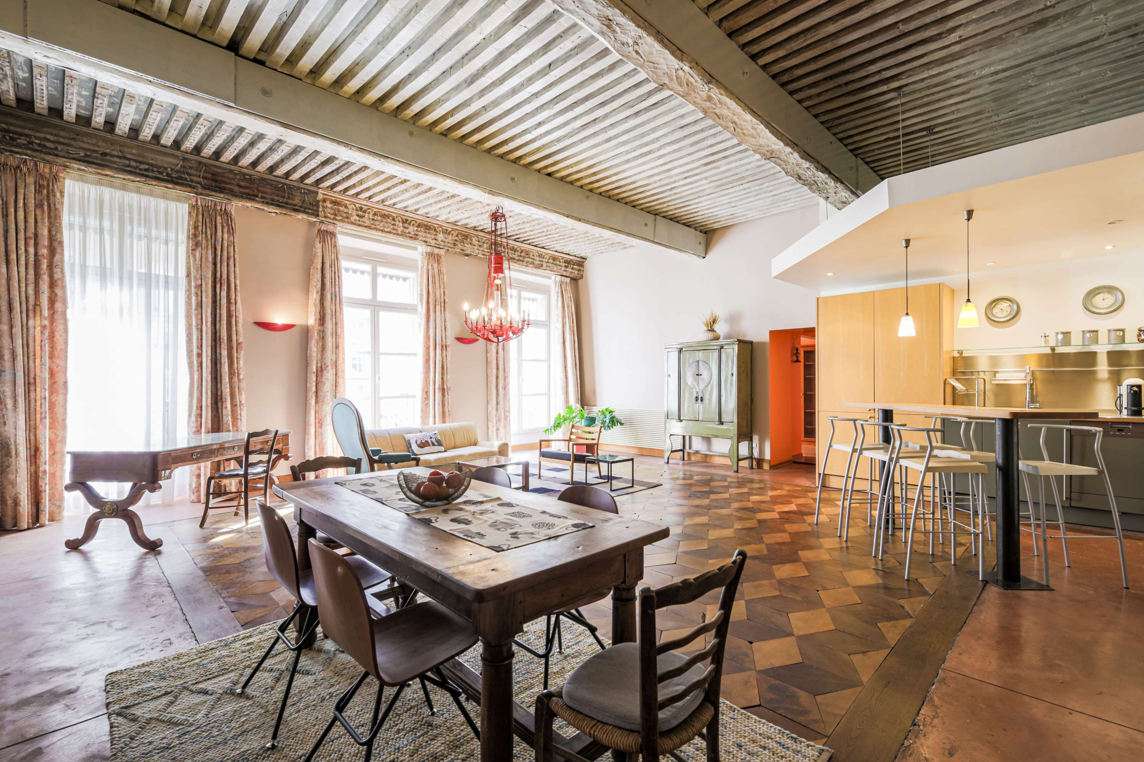 The image depicts a spacious, well-lit interior featuring a dining table, a lounge area, and a modern kitchen with bar seating.