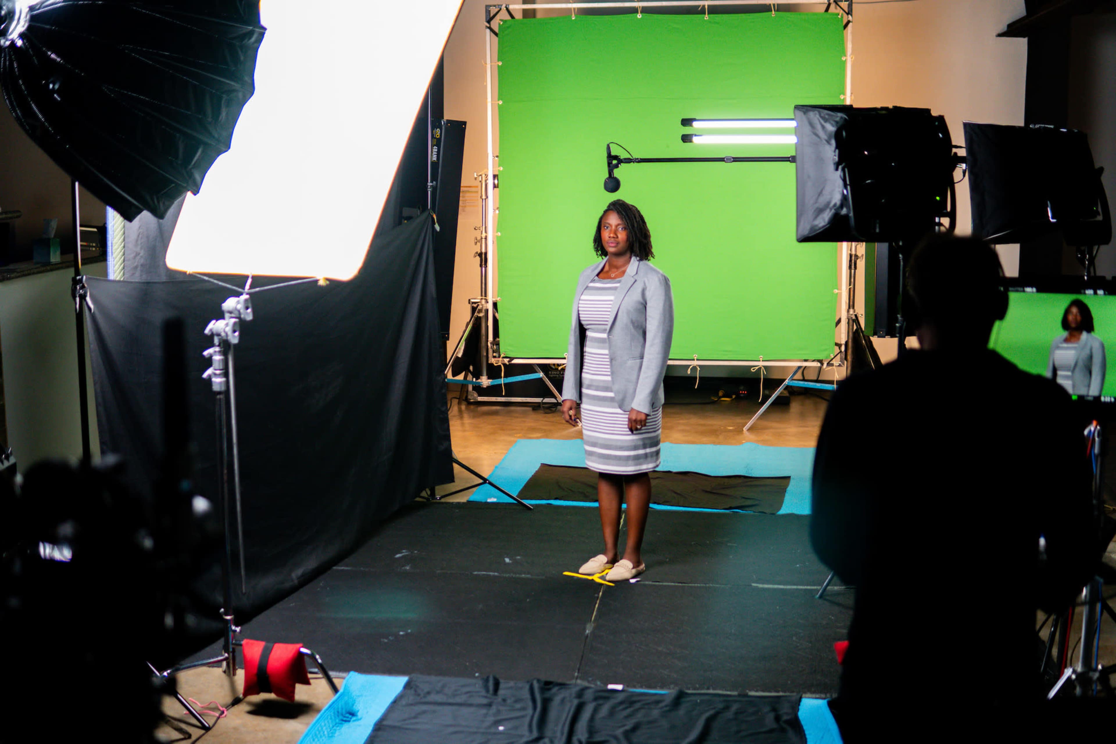 A woman stands on a black platform in front of a green screen, surrounded by studio lights and camera equipment.