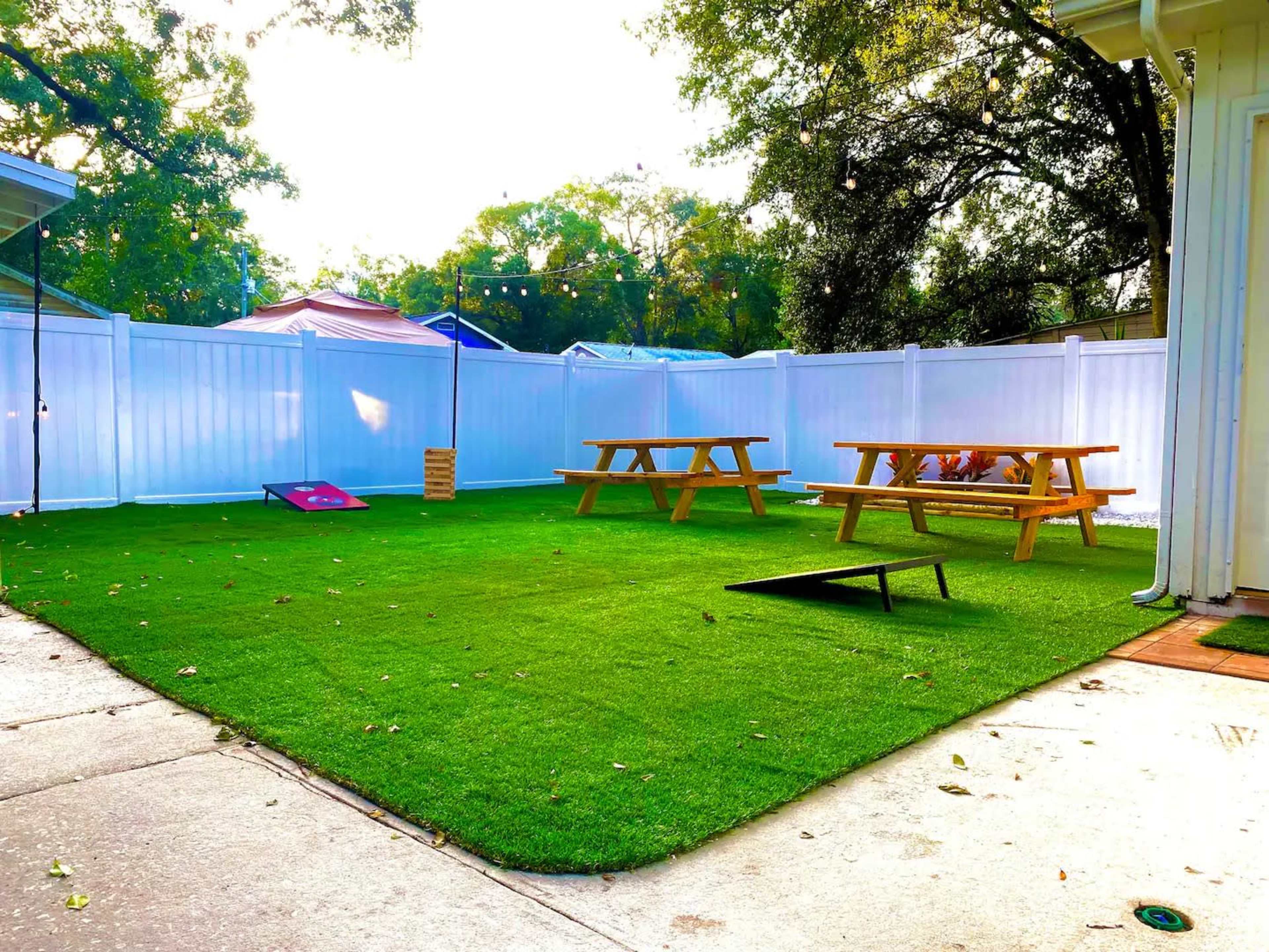 The scene features a backyard with artificial turf, two picnic tables, and a cornhole game set against a white fence.