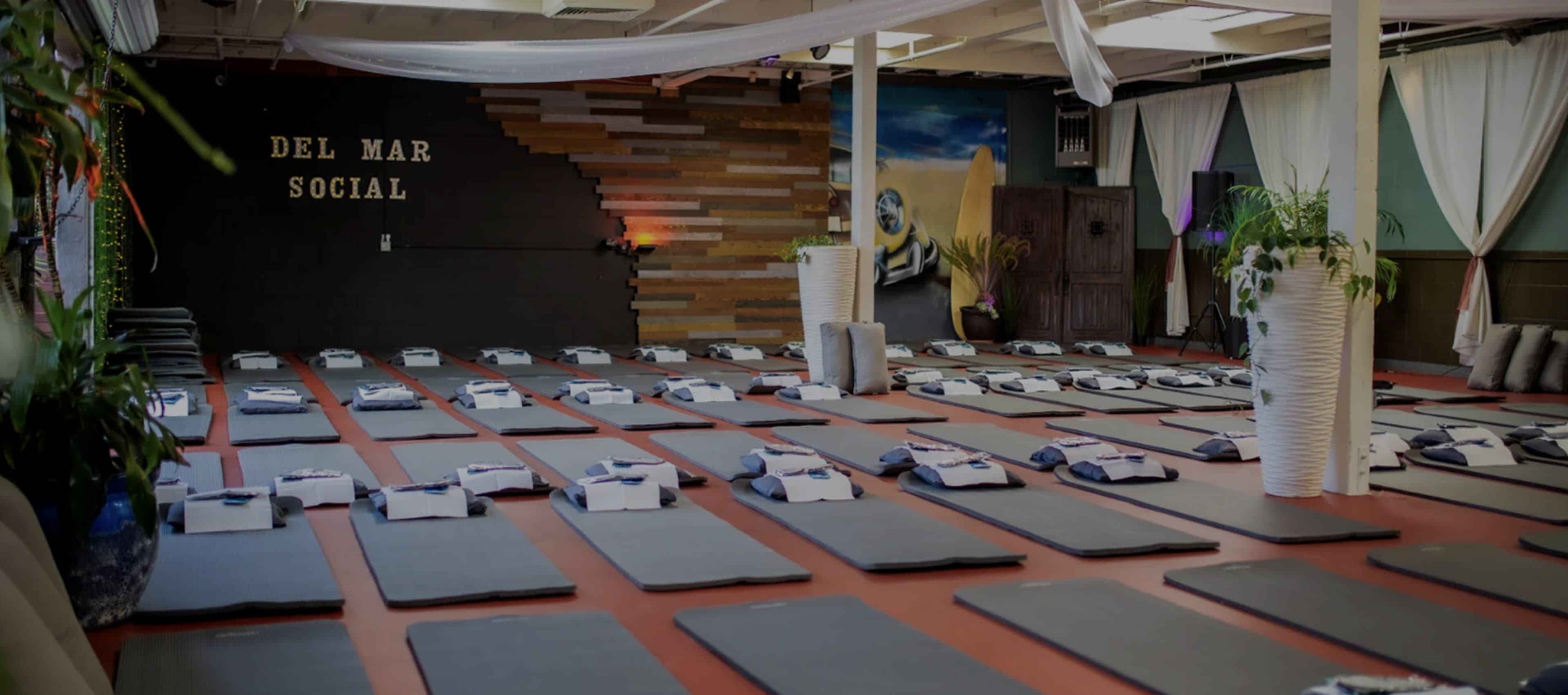 The image shows a spacious yoga studio with neatly arranged mats, plants, and a wooden feature wall with the words "DEL MAR SOCIAL" displayed prominently.