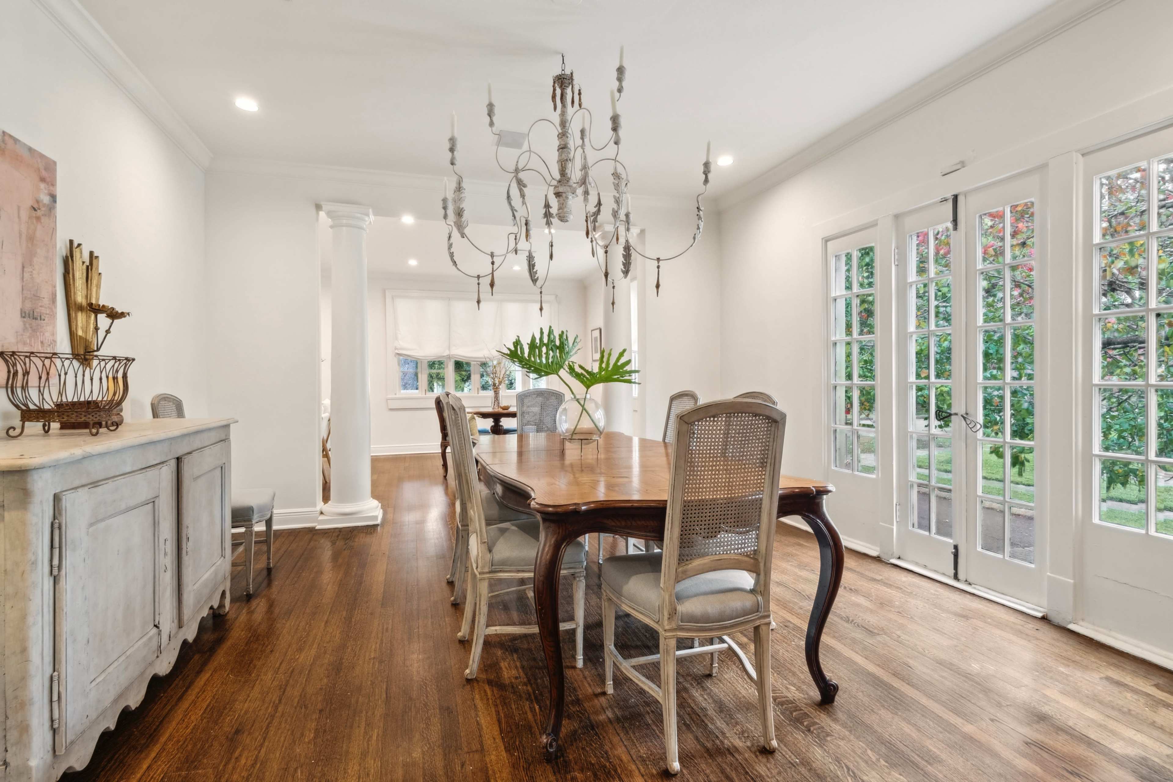 A spacious dining area features a wooden table surrounded by light-colored chairs, with a chandelier hanging above and large French doors leading to a garden.