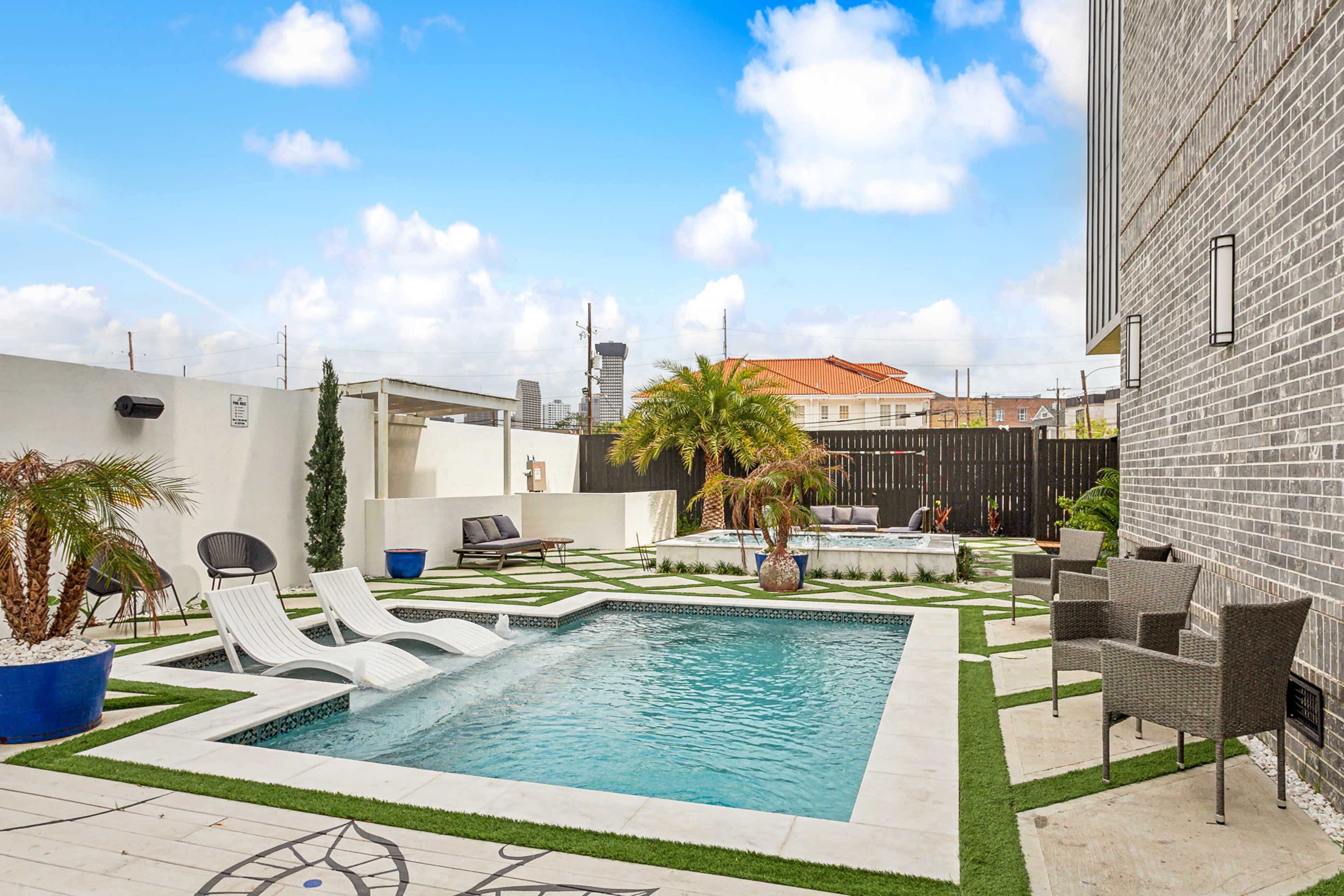 A swimming pool area with lounge chairs, potted plants, and outdoor seating surrounded by landscaped grass and stone paving.