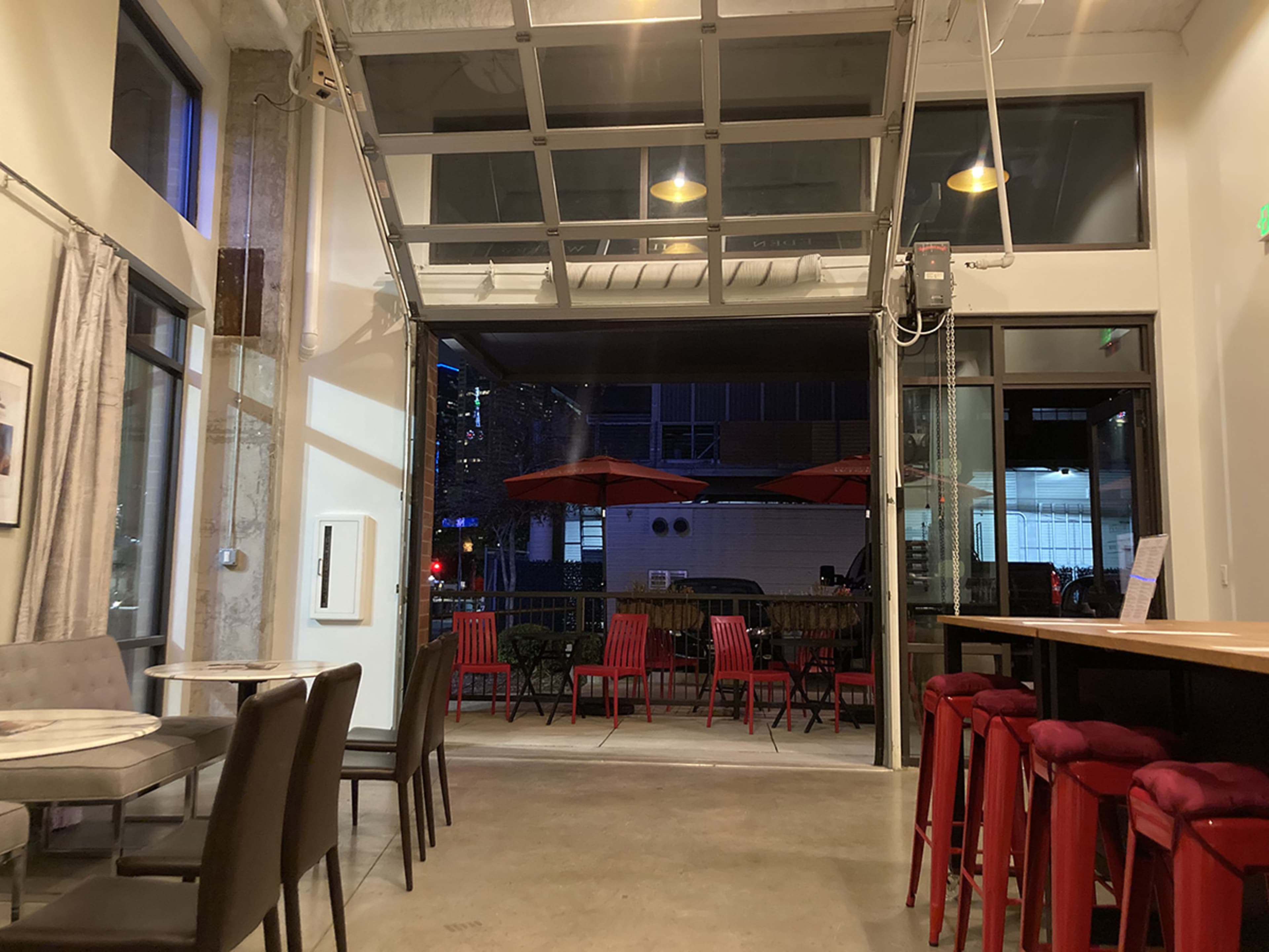 The image shows an interior space with a large open garage door that leads to an outdoor seating area, featuring red chairs and umbrellas.